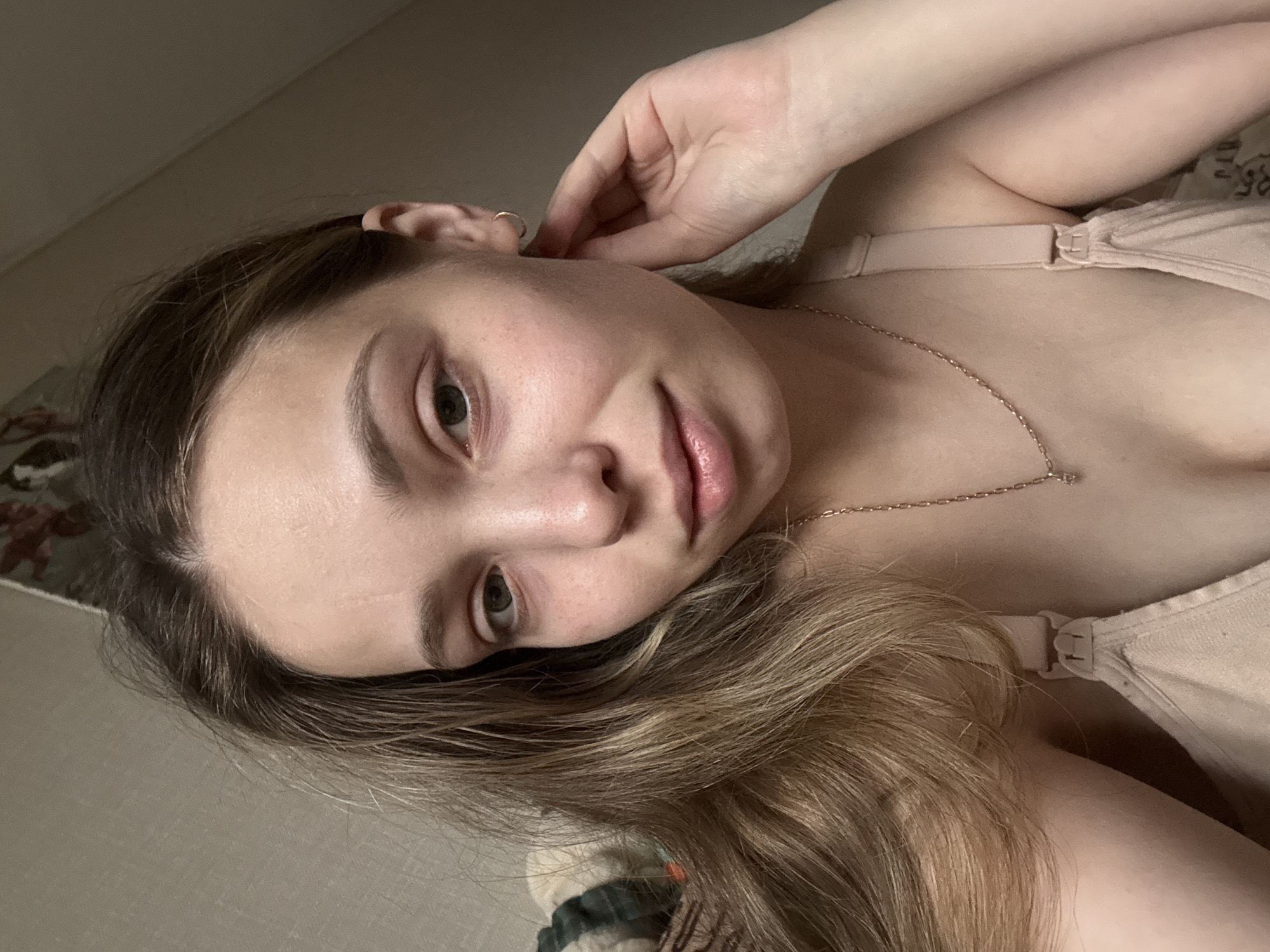This media file presents a close-up portrait of a young woman, captured from a high angle as she lies down and looks directly at the camera. The image is rotated 90 degrees clockwise. The woman, identified as being in Chaikovsky, Russia, has light-colored eyes (appearing green or hazel) and light brown hair with blonde highlights, which cascades around her. Her right hand is gently placed near her ear, drawing attention to a small hoop earring.
She wears a light beige or nude-colored bra or top, with a visible clasp on the strap over her right shoulder. A delicate gold-colored chain necklace with a small pendant is visible on her chest. The woman's expression is calm and serene, with her lips slightly parted. The background is softly blurred, revealing a light-colored wall or ceiling and patterned fabric, likely a blanket or bedspread. The lighting is soft and diffused, indicating an indoor setting. There is no discernible text within the image.