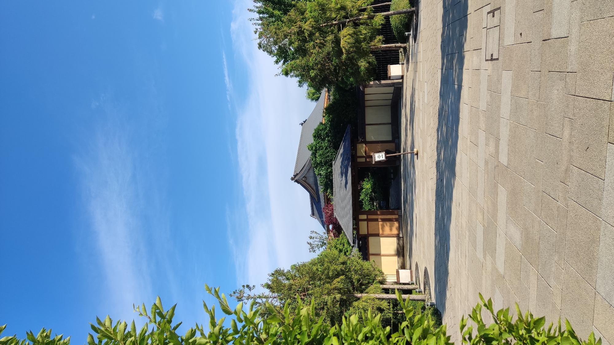 The image shows a paved courtyard leading to a traditional Japanese-style gate and building. The building has a dark, tiled roof with distinctive curves, and the walls are a light tan color. The gate is made of dark wood and is flanked by vertical wooden posts. Several coniferous trees and some deciduous trees with red foliage are visible in the background. The courtyard is paved with large, light-colored square tiles. The sky is bright blue with a few wispy white clouds. A small wooden signpost stands in the courtyard in front of the gate. The lighting suggests it is daytime, likely morning or late afternoon, as indicated by the shadows cast on the courtyard. The scene is tranquil and appears to be part of a garden or temple complex. There are no people or animals visible in the image. The location is not in Krasnodar, Russia, but rather appears to be in Japan, given the architectural style and landscaping.