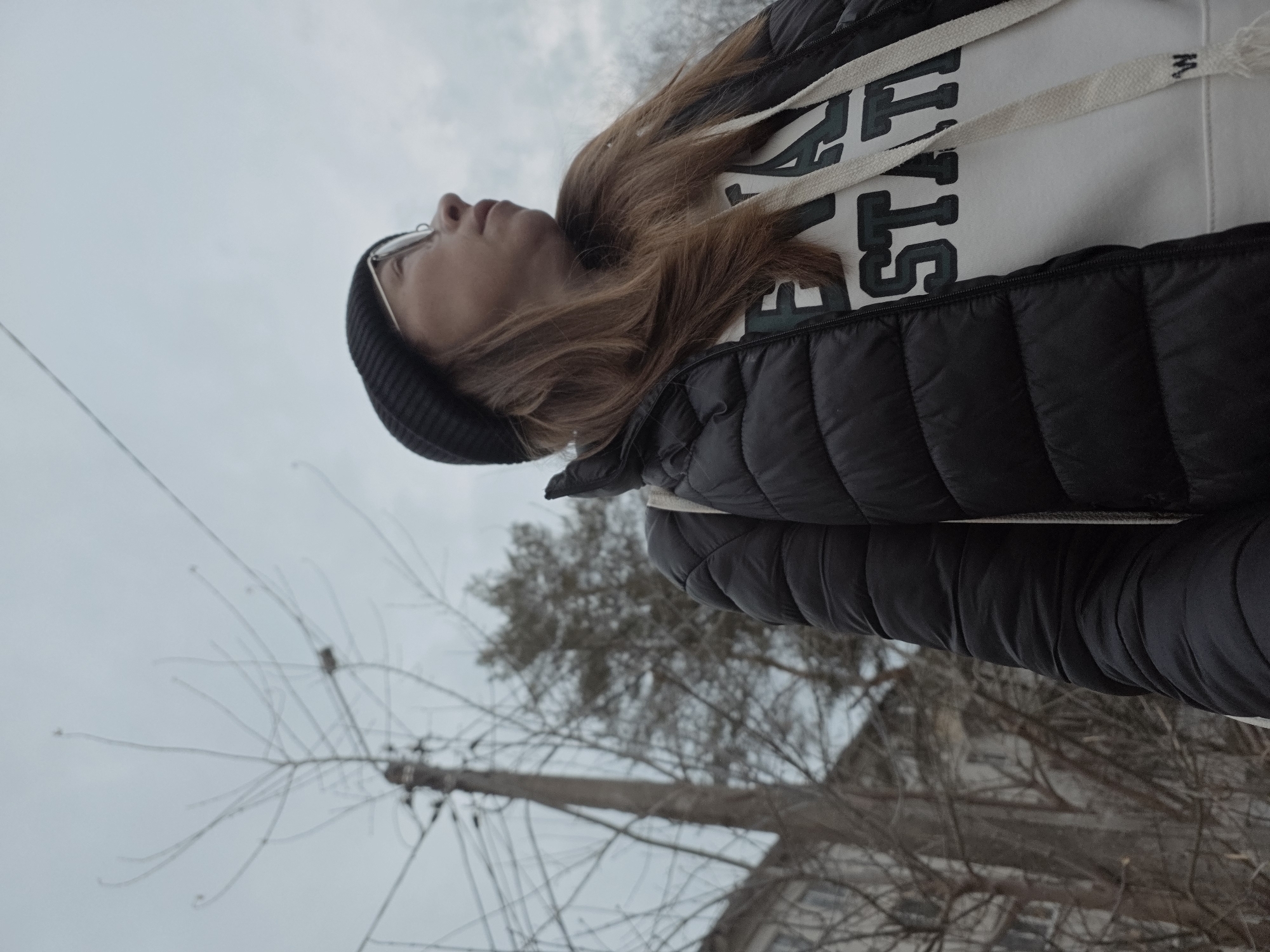 A woman wearing a black beanie, glasses, white sweatshirt with "STAT" printed on it, and a black puffer jacket stands outside, looking up. Bare trees and utility poles are visible against a grey sky. The location is the city of Chaikovsky, Russia. A building is partially visible at the bottom of the frame.
