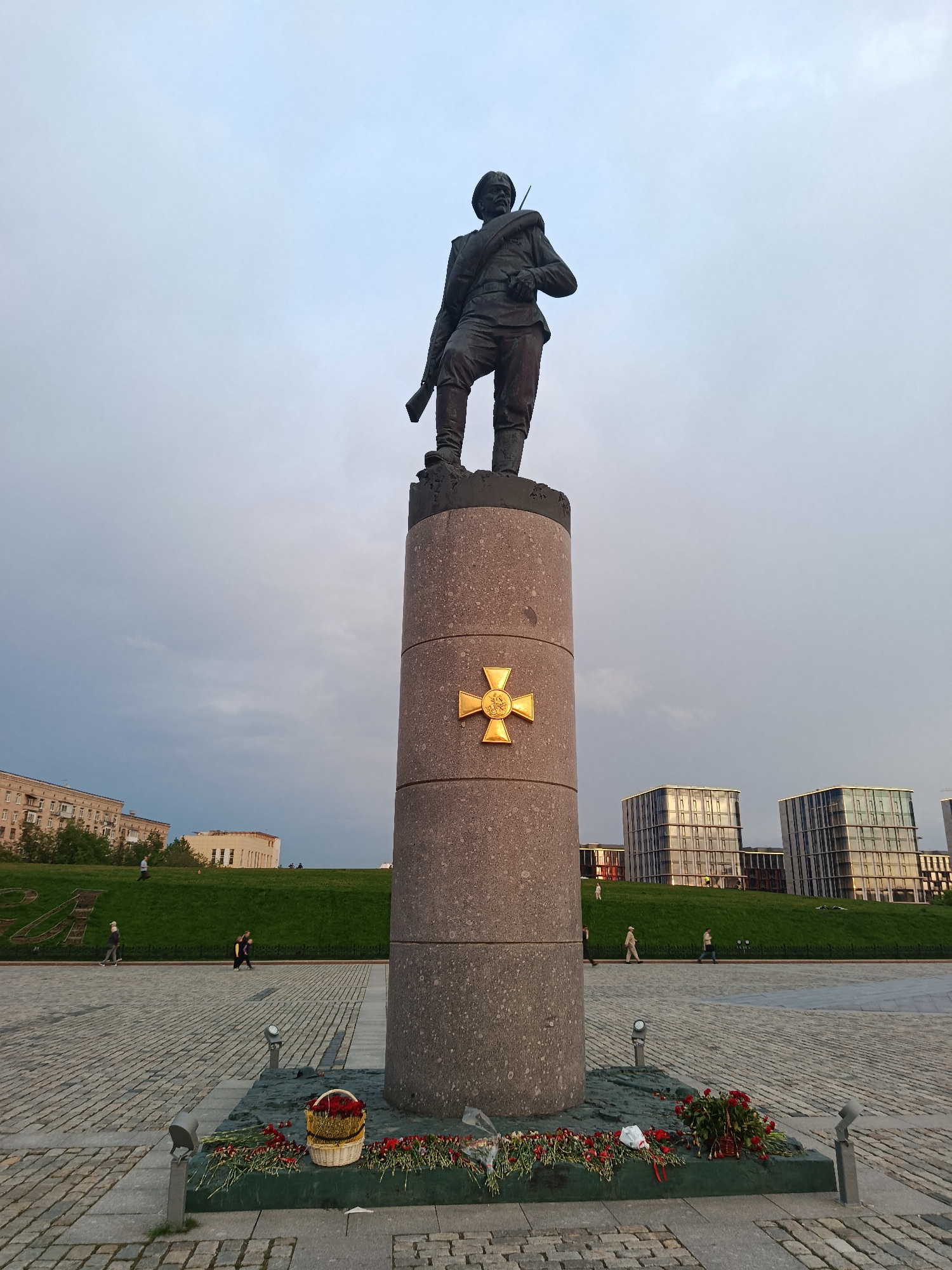 The image depicts a bronze statue of a soldier standing atop a cylindrical stone column in a public square. The statue is detailed, showing the soldier in uniform with a rifle. A gold-colored cross is affixed to the column. At the base of the monument, there are flowers and a basket of red roses. The square is paved with cobblestones, and several people are walking in the background. Beyond the square, there is a grassy slope with buildings visible in the distance. The buildings appear modern with glass facades. The sky is overcast. The location is likely a memorial site in Moscow, Russia, possibly dedicated to military personnel.