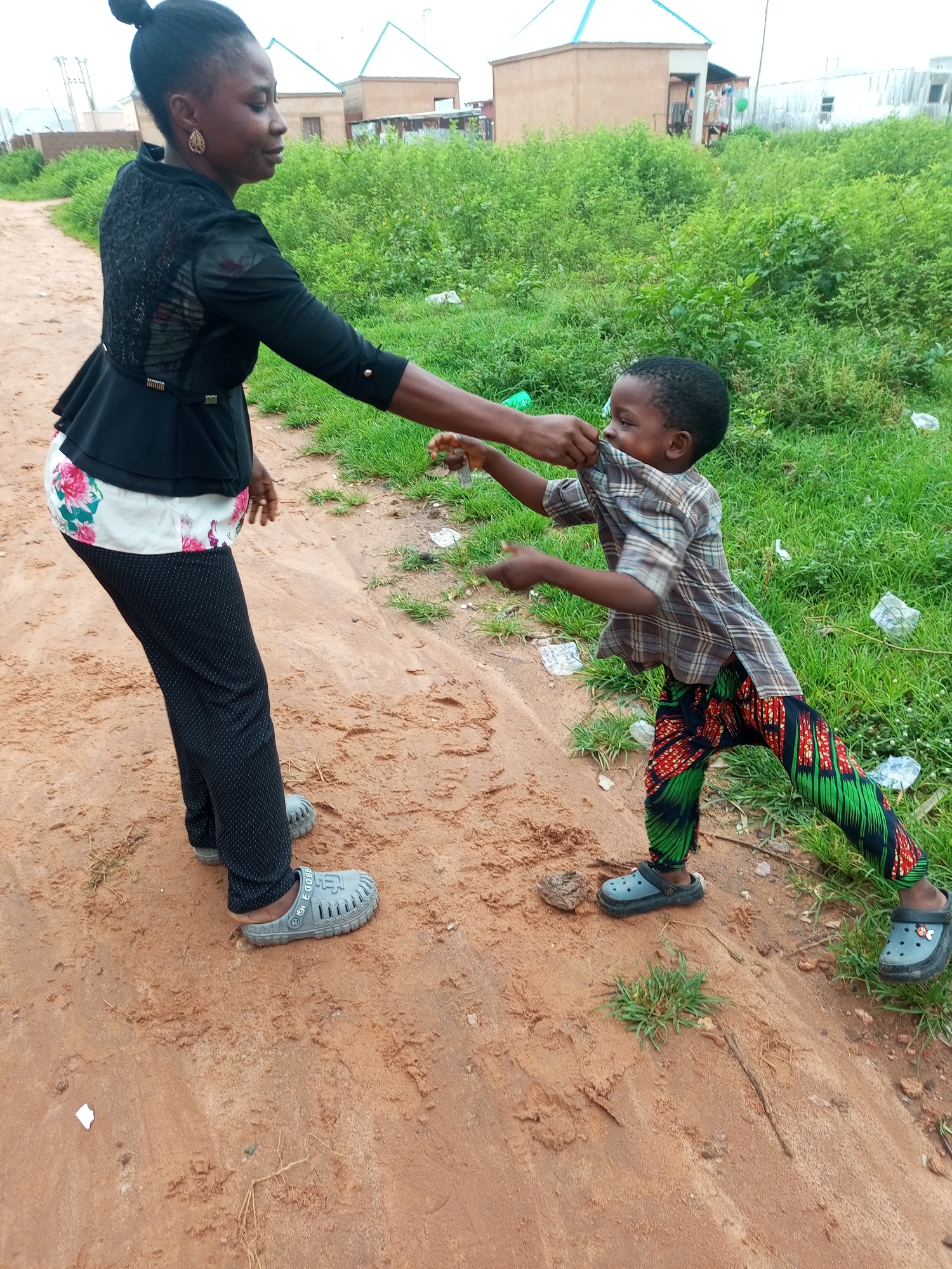 In Tempure, Nigeria, a woman and a young child are interacting on a dirt path adjacent to a grassy area. The woman, wearing a black ruffled top, a floral-patterned underskirt, and black patterned pants, stands on the left side of the frame. She has her hair pulled back in a bun and is wearing large hoop earrings. Her left hand is extended, and it appears she is holding something small, possibly food, which the child is trying to grasp.
The child, dressed in a plaid shirt and colorful patterned pants, is positioned to the right of the woman, facing her. He is actively pulling at the woman's shirt with both hands, seemingly in an attempt to get the item she holds. The child is wearing dark Croc-style shoes.
The environment is somewhat unkempt, with sparse green grass and scattered white plastic debris visible on the right side of the path. In the background, low-rise buildings with light-colored walls and light-blue roofs are visible, suggesting a residential area. The overall lighting indicates an overcast or cloudy day.