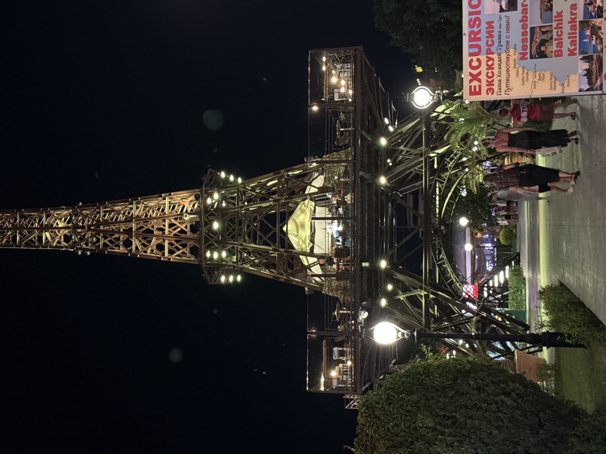The image shows a nighttime scene in the city of Varna, Bulgaria, featuring a tall, illuminated structure resembling the Eiffel Tower. The tower is made of metal scaffolding and has multiple levels with tables and chairs set up, suggesting it might house a restaurant or observation deck. Several people are visible walking on a paved path in front of the tower. To the right of the path, a signpost displays information about excursions, with text in both Cyrillic and Latin alphabets. The sign lists various locations such as "Nessebar", "Balchik", and "Kaliakra", which are popular tourist destinations along the Bulgarian Black Sea coast. The environment appears to be a promenade or a public area, with trees and planters adding to the ambiance. The lighting from the tower and streetlamps illuminates the scene, creating a warm and inviting atmosphere.