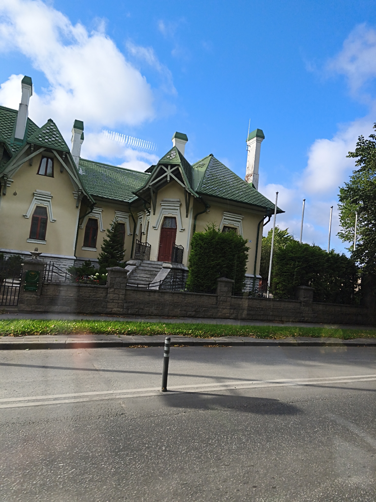 The image captures a section of the city of Tallinn, Estonia, showcasing a distinctive yellow house with a green tiled roof. The architecture features ornate trim around the windows and a prominent red front door. The house is bordered by a low stone wall, beyond which lies a paved road with white lane markings and a single dark bollard. A patch of green grass and some fallen leaves are visible between the wall and the road. In the background, several tall, thin flagpoles stand amidst dense green hedges and trees. The sky above is a vibrant blue, dotted with fluffy white clouds. The perspective suggests the photo was taken from a moving vehicle on the road, looking towards the house and its surroundings.