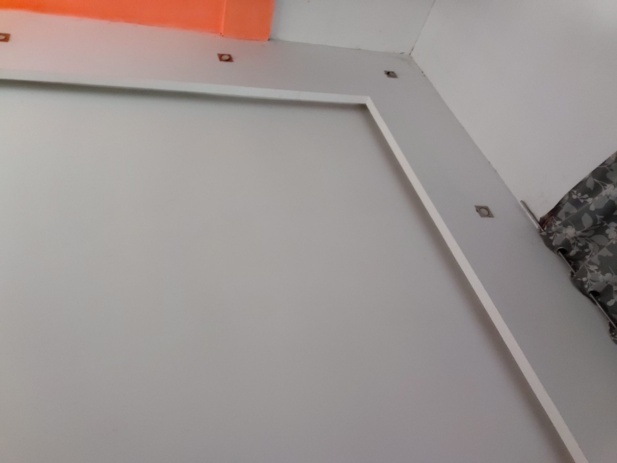 This image presents an interior architectural detail, likely a section of a room. A large, light gray, flat panel dominates the foreground, framed on its top and right edges by a thin white border. Along this white frame, and extending slightly onto the gray surface, are several small, square-shaped, brownish or coppery elements, each featuring a central circular indentation, possibly indicating recessed lights or mounting hardware.
Above the top white frame, a distinct, rectangular structure in a bright orange color is visible, adding a contrasting accent to the otherwise muted palette. In the upper right corner, a white ceiling and wall meet. To the far right, a patterned curtain or fabric is partially visible, displaying dark, possibly floral or leaf designs against a lighter background.
The photograph is taken from a low, upward-angled perspective, emphasizing the geometry of the walls and ceiling. The lighting is soft and even, suggesting an indoor environment without harsh shadows. No individuals or specific activities are depicted. The scene is static, focusing on the structural and decorative elements of the interior. No text is visible in the image. The provided context indicates the location is Santa Ana, Venezuela.