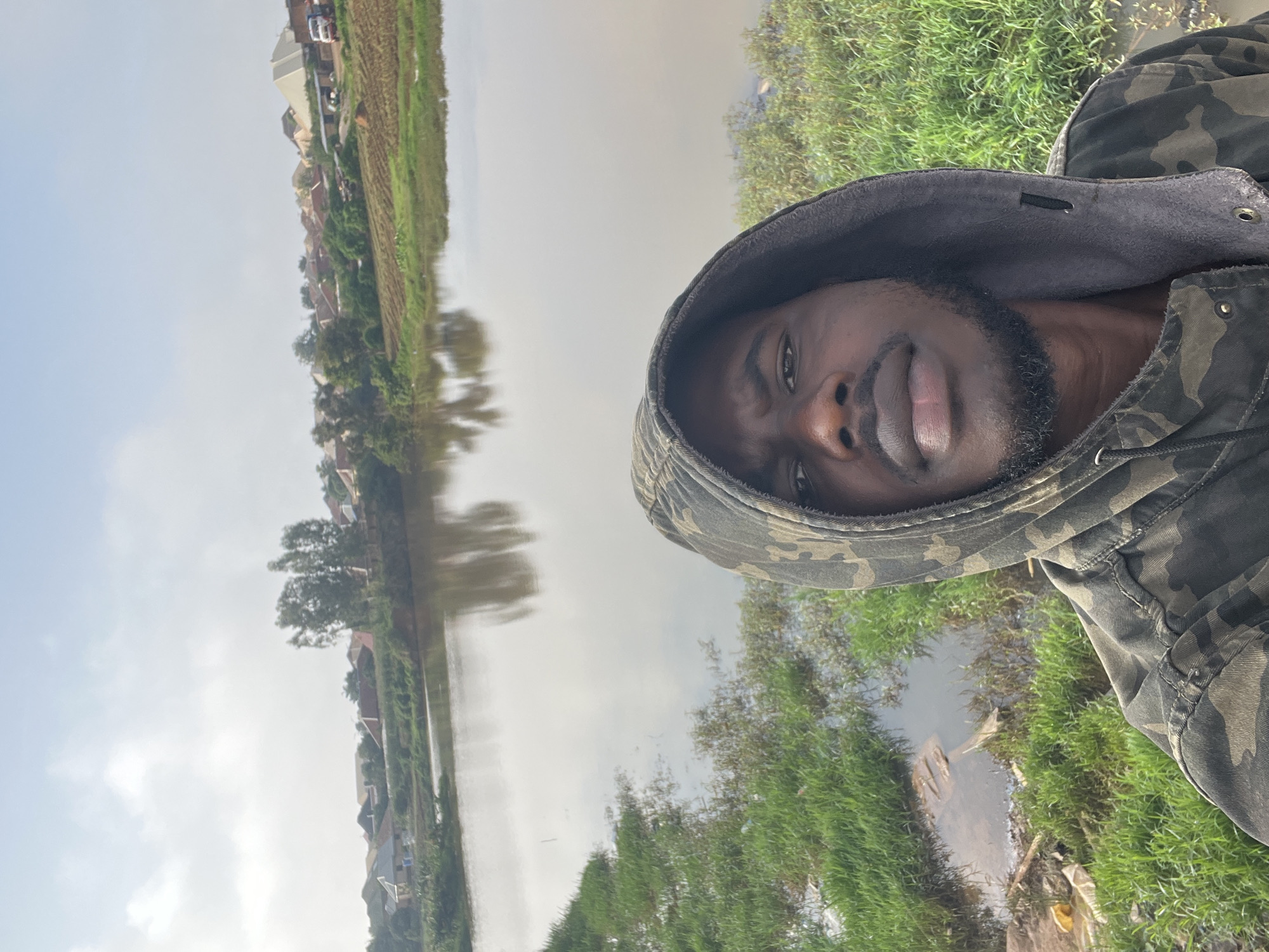 A dark-skinned man in a camouflage hoodie stands in the foreground, taking a selfie. He has a beard and is looking directly at the camera. Behind him, a body of water reflects the cloudy sky. The water's edge is lined with green grass and some debris. In the background, a residential area with houses and trees is visible, suggesting a semi-urban or suburban setting. The overall scene depicts a casual outdoor moment near a body of water, possibly in the city of Jos, Nigeria, given the architecture and vegetation.