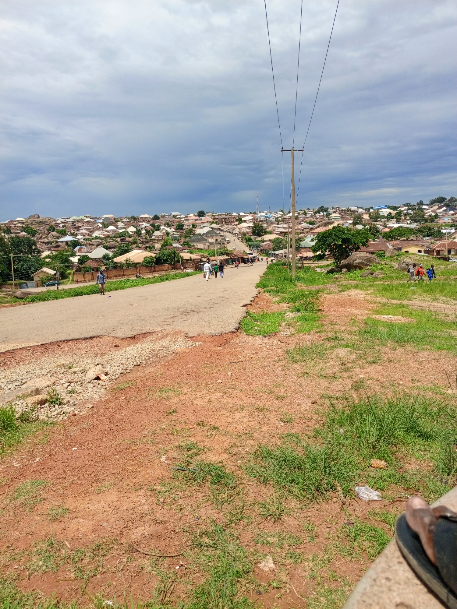 The image depicts a view of a town located on a hillside in Jos, Nigeria. The town is characterized by numerous buildings with corrugated iron roofs, clustered together on uneven terrain. A paved road runs through the foreground, curving towards the left and disappearing into the distance. Several individuals are visible walking along the road and on the grassy slopes. Power lines traverse the upper portion of the image, supported by a wooden pole. The sky is overcast with a mix of clouds. The foreground consists of reddish-brown soil with patches of green grass.