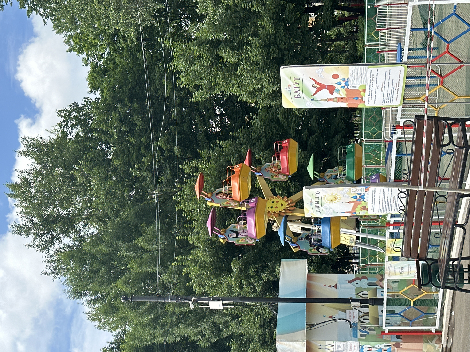 In Saransk, Russia, on a sunny day with scattered clouds, an amusement park scene unfolds. A small Ferris wheel, painted in vibrant yellow, pink, and orange, stands as the central attraction, its colorful gondolas occupied by unseen children. To the left, a whimsical mural depicts a fairy-tale cityscape with castles and towers. In the foreground, a wooden bench sits empty, facing the attractions. Two signs provide information about the park's features: one, titled "СОЛНЫШКО" (Solnyshko), features a smiling sun and a depiction of Moscow's St. Basil's Cathedral, while the other, "БАТУТ" (Batut), showcases a leaping figure above a similar Russian architectural scene. A green trash bin is tucked behind the bench. The park is enclosed by a colorful fence, and the background is dominated by lush, green trees.