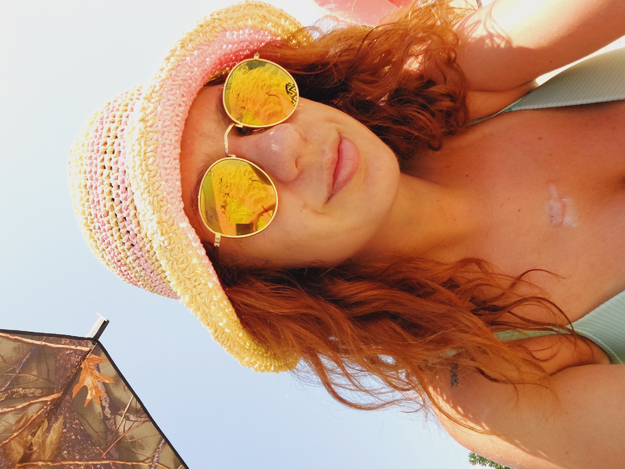 A young woman with auburn hair and fair skin is shown from the chest up, looking towards the camera. She is wearing a straw hat with pink and yellow accents and reflective sunglasses. The background is a clear blue sky, with a portion of a camouflage umbrella visible in the upper left corner. The woman appears to be outdoors, likely on a beach or in a sunny location, given the attire and lighting. Her left shoulder has a small tattoo, and a scar is visible on her chest. The image is a selfie, indicating the woman is taking the photograph herself. There is no information to place the scene in Sozopol, Bulgaria.