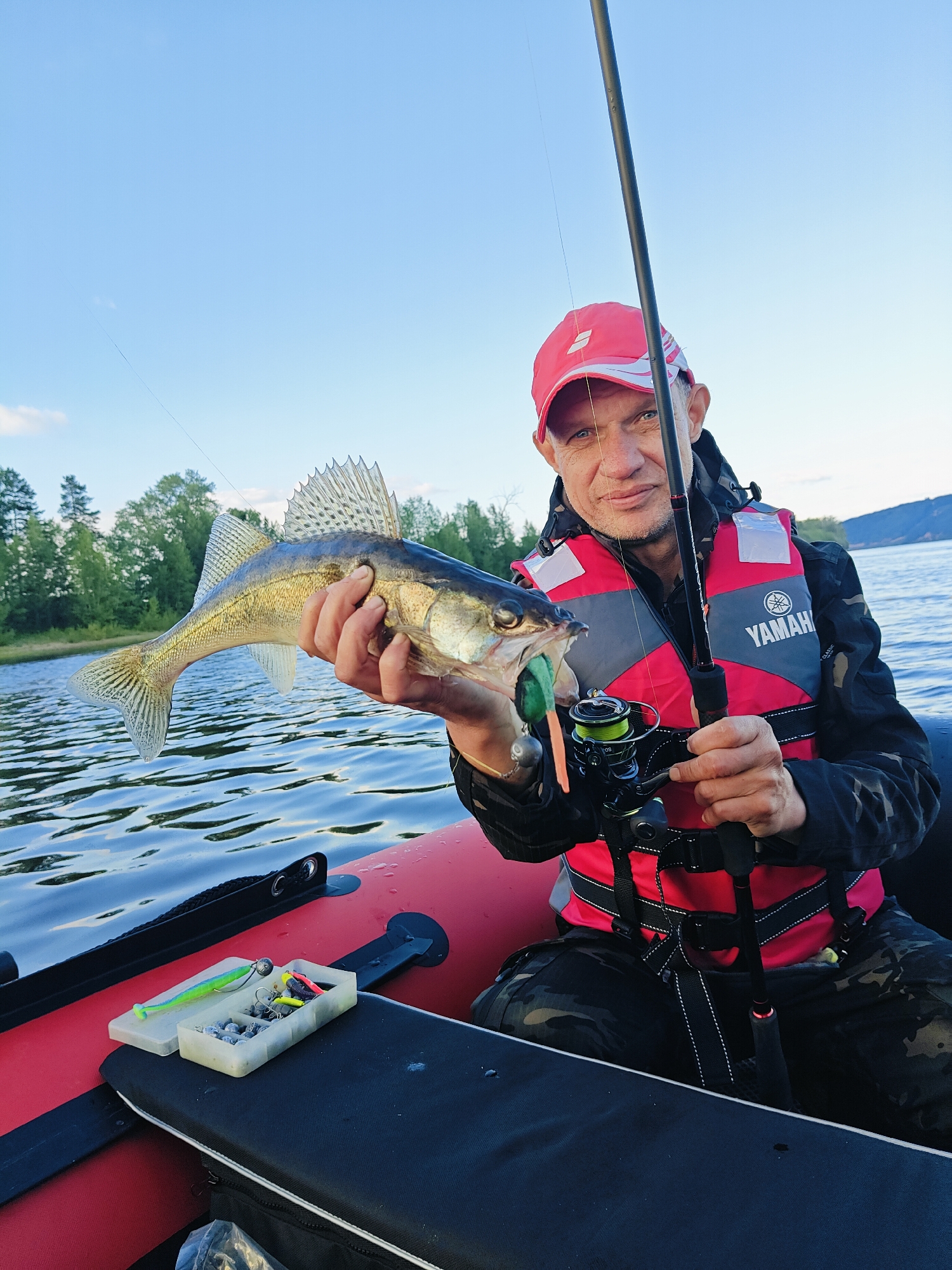 A man in a red baseball cap and a red life vest with "YAMAHA" printed on it is sitting in a red inflatable boat. He is holding a freshly caught fish in his left hand and a fishing rod in his right hand. The fish is a walleye, with its mouth open, showing a green lure. The man is smiling and looking at the camera. The boat is on a body of water, with trees and hills in the background. The sky is blue with some clouds. There is a tackle box with fishing lures on the boat. The scene appears to be set in the late afternoon or early evening, with the sun casting a warm light. The overall mood is one of satisfaction and enjoyment of fishing. The location is Ol'khovskoye, Russia.