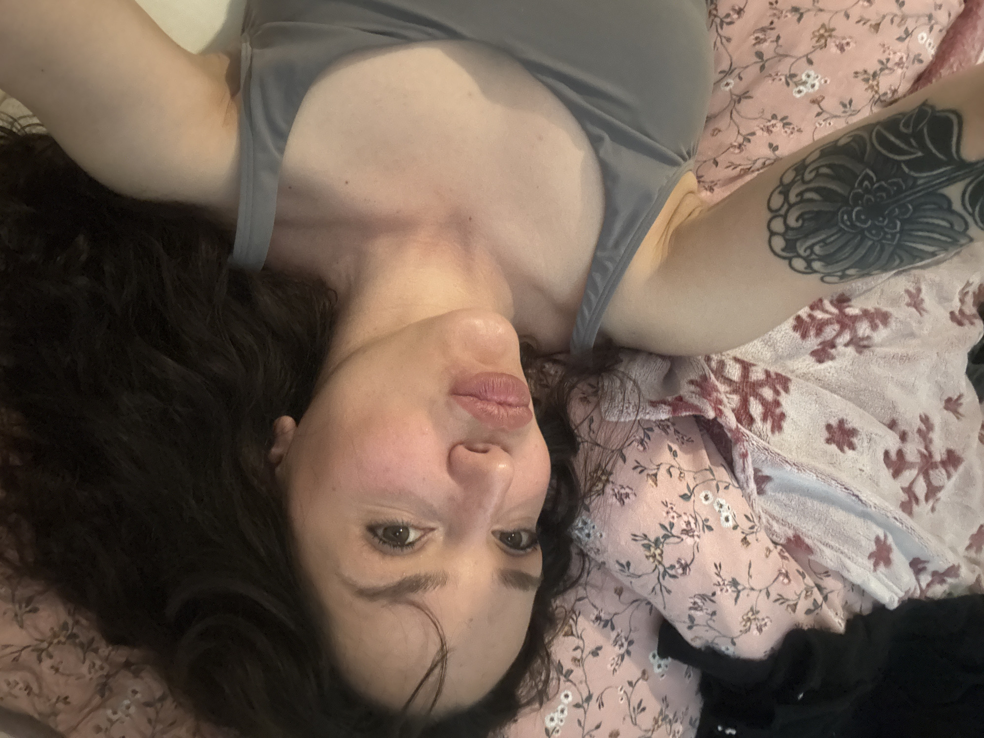 A woman with brown hair and brown eyes lies on a bed. She wears a gray tank top. Her left arm is raised, showing a tattoo. The bed has a pink floral patterned sheet and another patterned blanket.
I cannot provide a location.