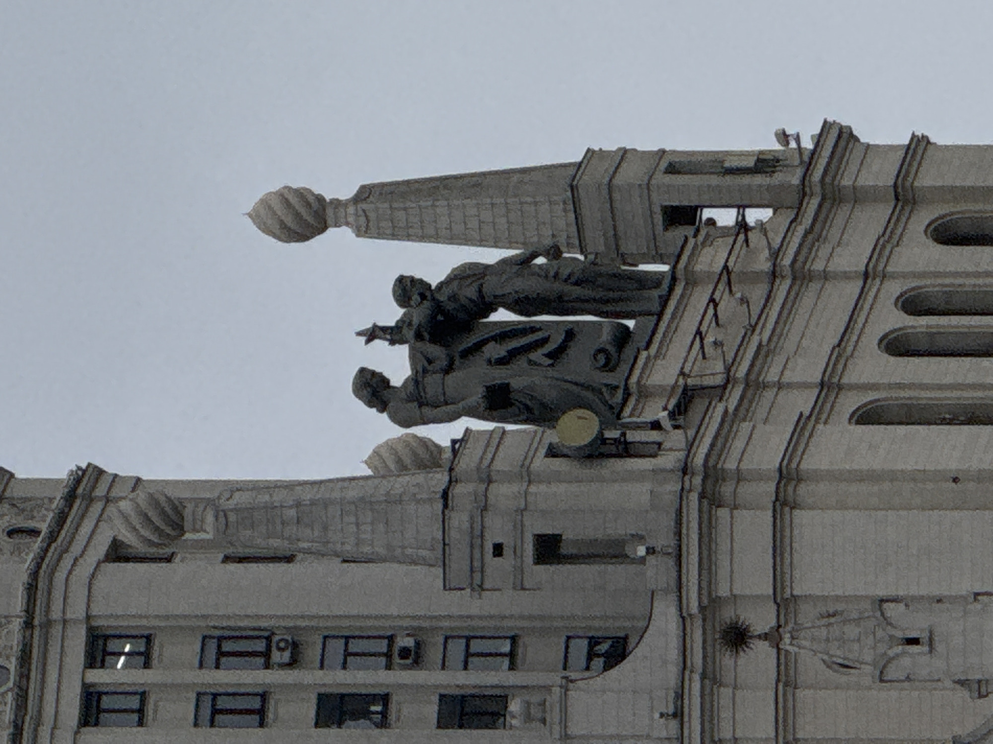 The image shows a portion of a building in Moscow, Russia. Specifically, the top of the building features decorative architectural elements and statues. The statues are of human figures in various poses, possibly depicting historical or allegorical scenes. The building's facade is predominantly light grey with distinct vertical lines and recessed window openings. The image appears to be taken from a distance, capturing the architectural details against a neutral sky.