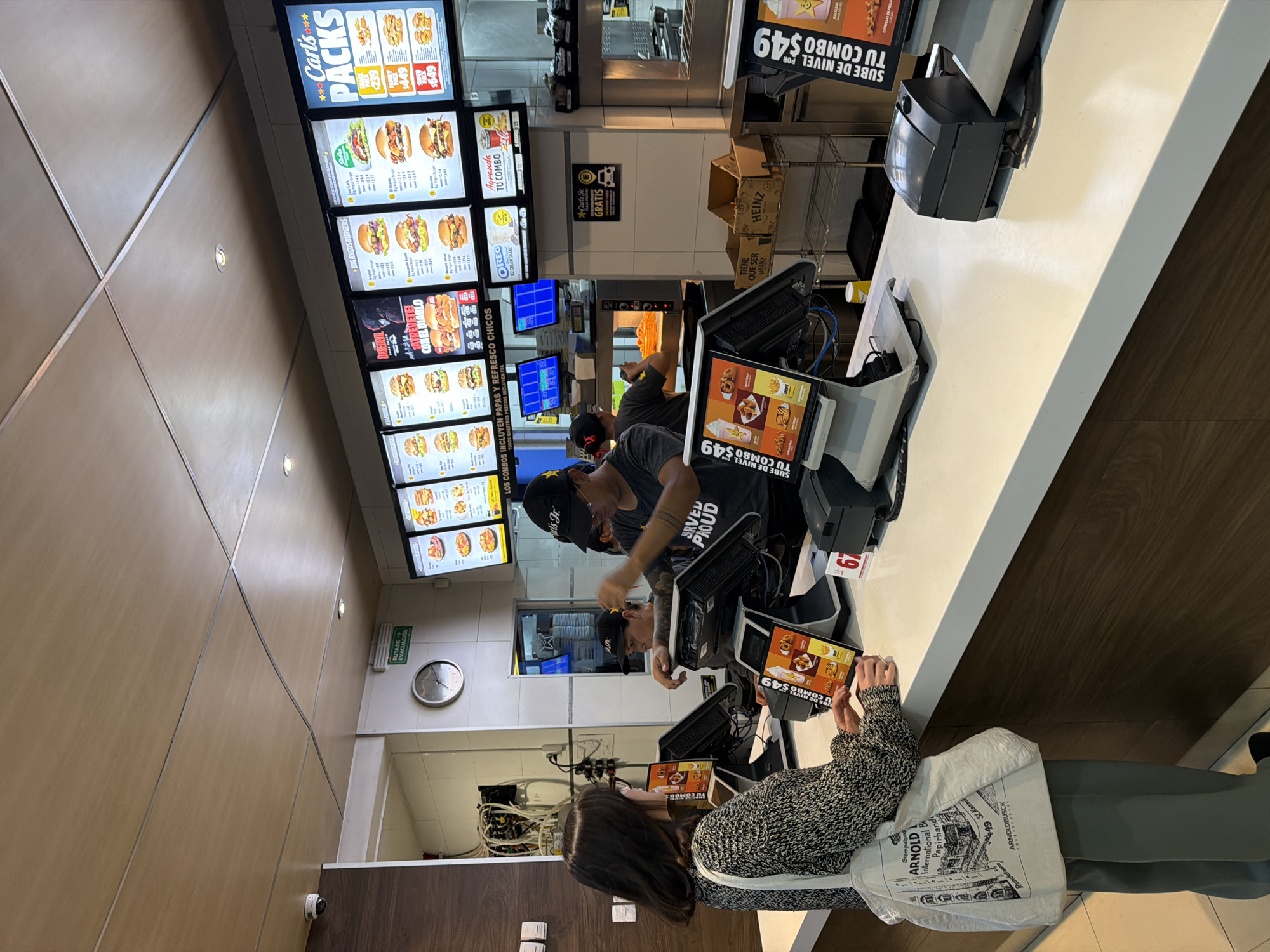 This image depicts the interior of a Carl's Jr. fast-food restaurant located in San Bernardino Tlaxcalancingo, Mexico, specifically at the order counter and adjacent kitchen area. The scene is brightly illuminated by overhead lighting and illuminated menu boards.
In the foreground, a female customer with dark, shoulder-length hair and glasses stands facing the counter. She wears a patterned grey and white sweater over green pants and carries a white canvas tote bag with a black architectural drawing and text including "ARNOLD INTERNATIONAL" and "ARNOLDBUSCH." Her right hand is positioned over a touchscreen order terminal, displaying a Carl's Jr. combo offer.
Behind the counter, two male employees are visible. The employee closer to the camera wears a black baseball cap with a yellow star and a dark grey t-shirt featuring "SERVED PROUD" text and "Carl's Jr." on the sleeve. He has visible tattoos on his right arm and is positioned to interact with customers, with his left hand near a monitor and his right arm partially raised. A second male employee, also wearing a black cap, is partially visible further back, actively working at a food preparation station within the kitchen, where a fryer or oven containing orange food items is apparent.
The white counter surface extends across the image. Three black and grey touchscreen order terminals are visible on the customer side, with the customer interacting with the leftmost one. Above the counter, a series of large, illuminated digital menu boards display various Carl's Jr. food items, including "Big Angus Burgers," "Carl's Packs," and promotional images. One board highlights "Daredevil" and "ATREVETE AL DIABLO." Additional monitors are visible on the employee side of the counter.
The background reveals a clean kitchen area with stainless steel elements and shelving units, one containing cardboard boxes, one of which is marked "HEINZ." A white tiled wall features a circular analog clock displaying approximately 1:10 and a green "RUTA DE EVACUACION" (Evacuation Route) sign. Wires and electrical conduits are exposed on the left wall. The ceiling consists of light brown acoustic tiles with recessed lighting fixtures. A white dome security camera is mounted in the upper left corner of the ceiling.