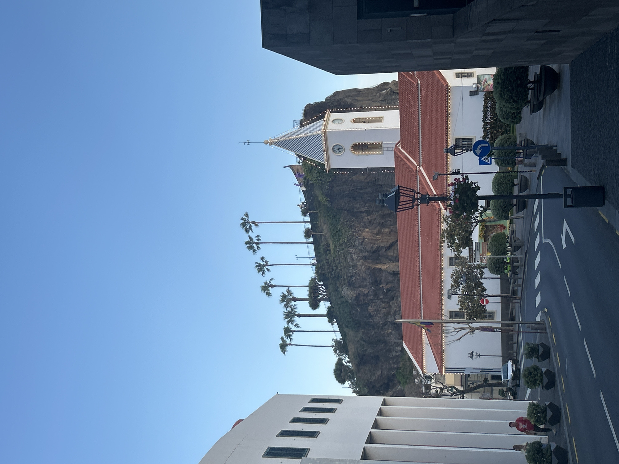 The image shows a view in Câmara de Lobos, Portugal. A white church with a blue and white checkered spire sits atop a rocky cliff, with palm trees lining the ascent. Buildings frame the foreground, one white and one gray. A road with white stripes runs alongside the base of the cliff, and street lamps and small trees line the sidewalk. Two people are visible near the bottom right of the frame. The sky is a clear blue.
