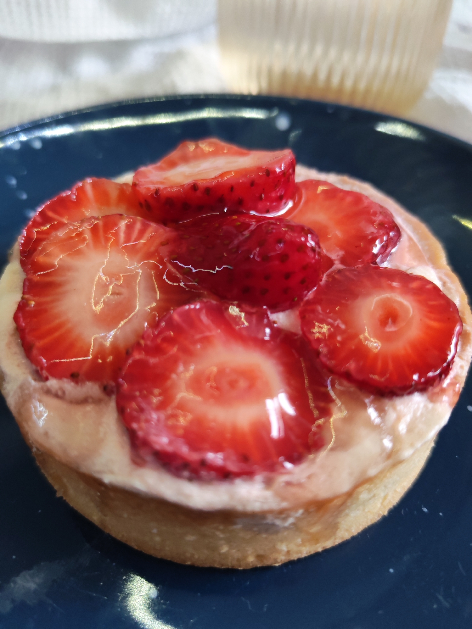 A close-up shot shows a strawberry tart on a dark blue plate. The tart has a golden-brown crust filled with a light-colored cream and topped with sliced fresh strawberries glazed with a clear, shiny topping. The background includes a blurry white cloth and a translucent glass. The image appears to be taken indoors with natural lighting. The scene is located in the city of Izhevsk, Russia.