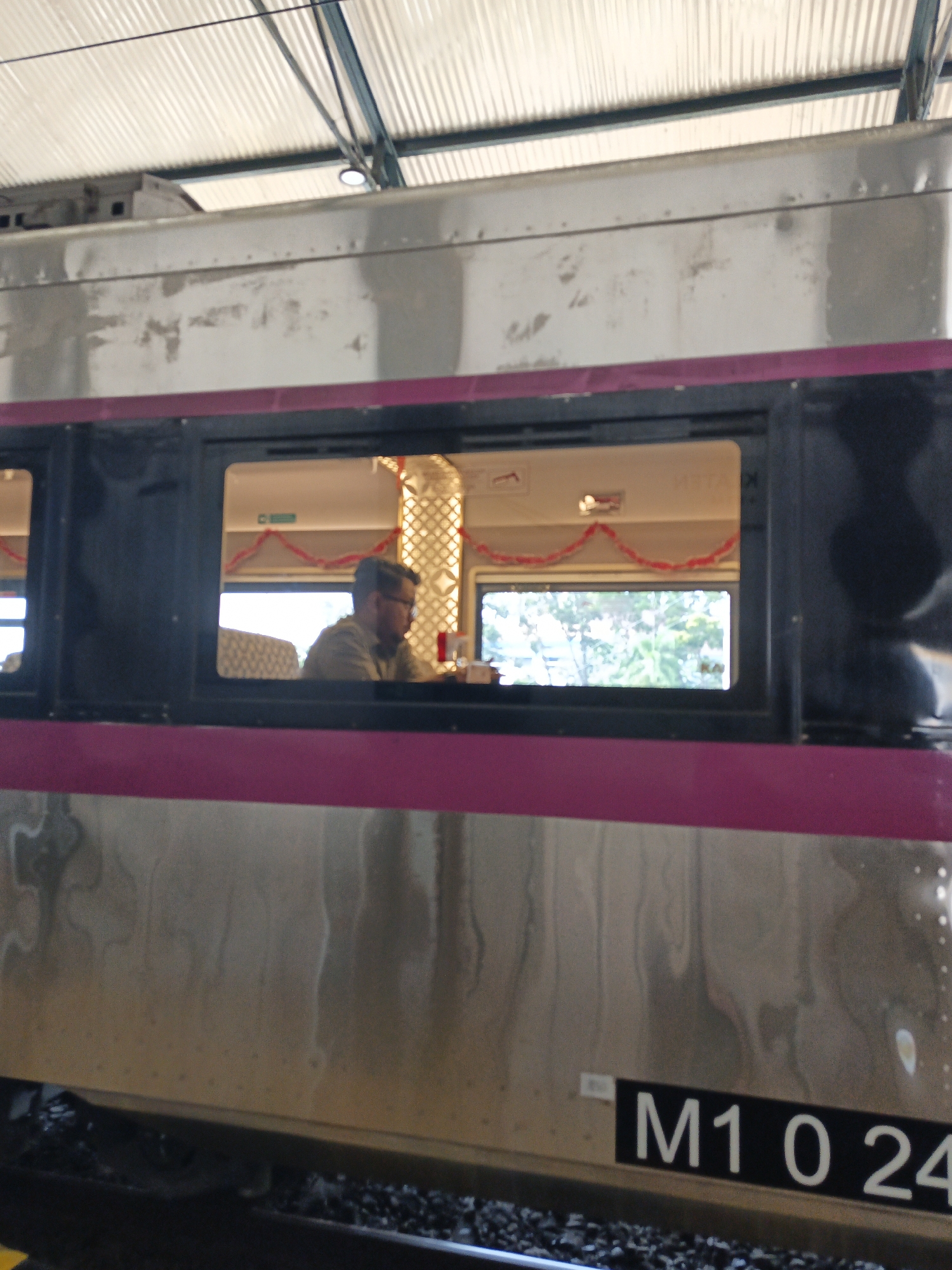 The image captures a view of a train from the outside, focusing on a window that reveals the interior. Inside the train, a man wearing glasses and a collared shirt is visible, seated at a table. The interior is decorated with red garland, suggesting a festive or seasonal theme. Beyond the man's window, another window shows a blurred view of trees and greenery outside, indicating the train is in motion or near a natural environment. The exterior of the train is metallic with a magenta stripe running horizontally along the windows. At the bottom of the train car, a black plate with white text reads "M1 0 24". The overall scene suggests a transportation setting, likely a train station or a moving train. The prompt mentions the location of Klaten, Indonesia, but there are no specific visual cues within the image that definitively confirm this location.