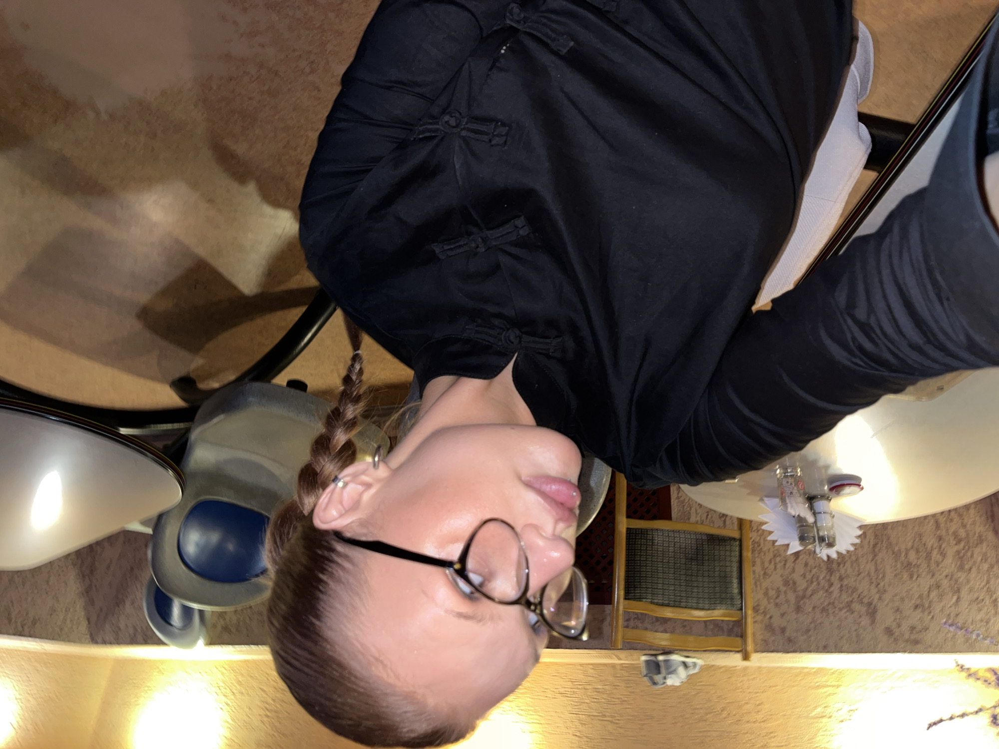 A woman takes a selfie in an indoor dining establishment located in the city of Chaikovsky, Russia. She is positioned in the mid-shot, facing left with a slight upward gaze. She has fair skin, wears black-rimmed glasses, and has a small silver labret piercing on her lower lip. Her hair is pulled back into two visible braids, and she wears a silver hoop earring in her right earlobe along with a helix piercing. She is dressed in a black tunic-style top featuring traditional Chinese frog closures. To her immediate left is a round white table set with a salt shaker, pepper shaker, a stack of white paper napkins, a small red object, and a clear glass. In the background, behind the woman, are various chairs including a wooden chair with a dark patterned backrest and a grey and blue upholstered chair. The walls feature textured, light brown paneling, illuminated by warm, yellow wall-mounted lights. The floor appears to be a light brown tiled surface.