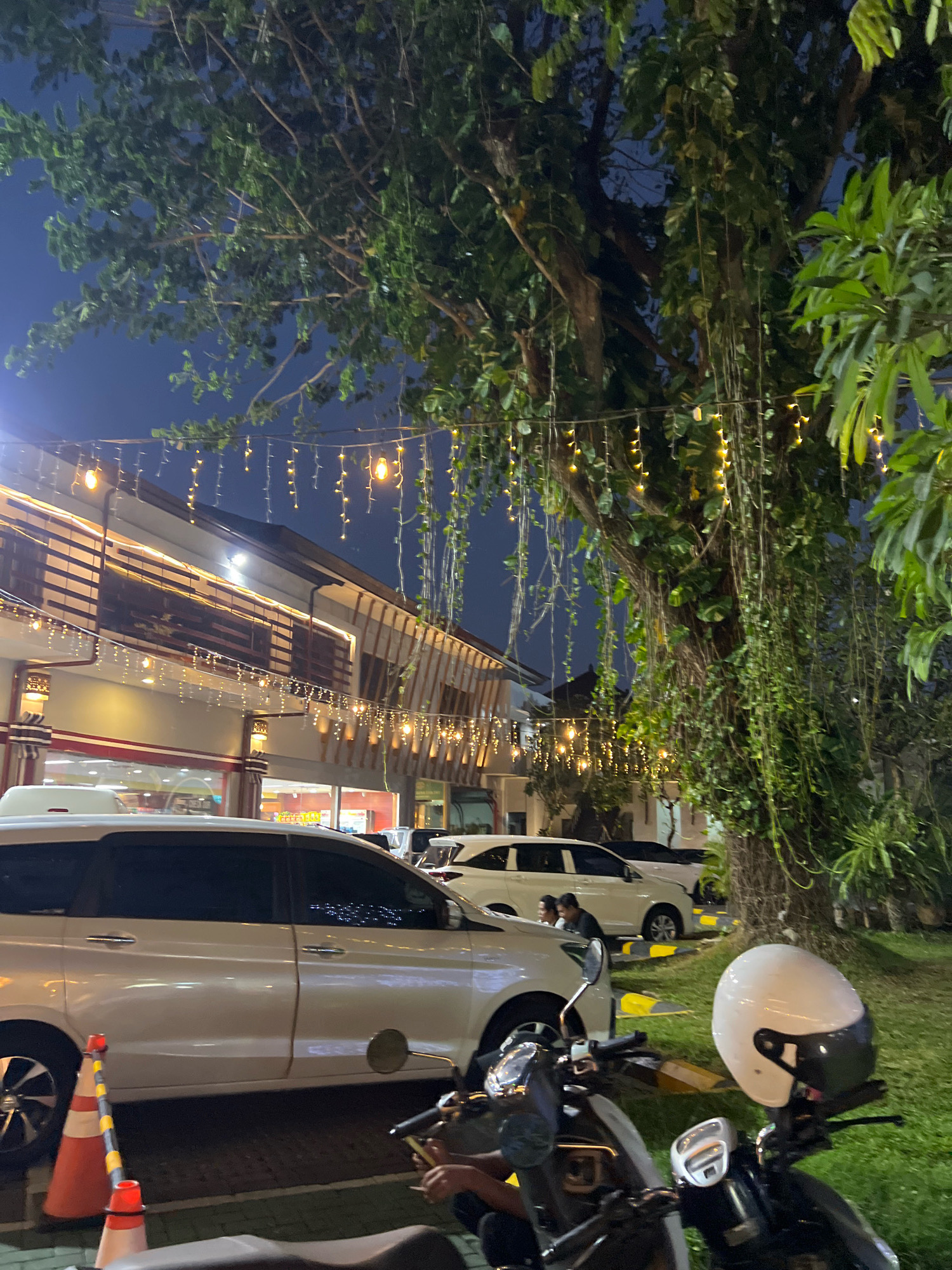 The image shows a street scene at night in Kuta, Indonesia. A white MPV is parked in front of a building adorned with string lights. A motorcycle is parked in the foreground, with a white helmet placed on its handlebar. The building appears to be a restaurant or shop, with the interior visible through large windows. The street is paved, and there is a patch of grass to the right, with a large tree covered in vines. The sky is dark blue, indicating it is nighttime. The overall atmosphere is festive, likely due to the decorative lights.