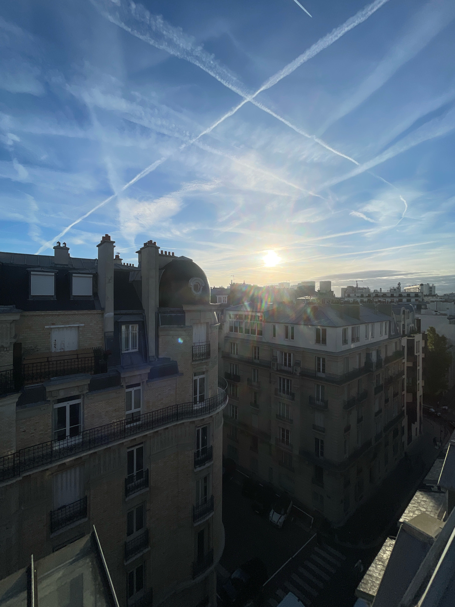 This is a high-angle, outdoor, daytime, wide shot taken from a building in Paris, France, looking out over a cityscape. The sky is a bright blue, with many white contrails crisscrossing from airplanes, forming an "X" pattern in the upper portion of the sky. The sun is visible in the upper center of the frame, slightly to the right, with lens flare emanating from it.
In the foreground and midground are several multi-story apartment buildings with mansard roofs and ornate facades. The building on the left is made of light-colored brick with dark grey trim around the windows and balconies. It features numerous windows, many with white shutters. A curved balcony with black railings is visible on the right side of this building. The building in the midground is similar in style and height, also with light-colored brickwork and a mansard roof. This building has a prominent curved corner.
In the background, more buildings of varying heights are visible, some with cranes suggesting ongoing construction. The streets below are dark, with a few cars visible. A crosswalk is discernible on the street in the lower right quadrant of the image. No people are clearly visible. The overall scene depicts a typical urban environment in Paris during what appears to be sunrise or sunset, given the lighting conditions.