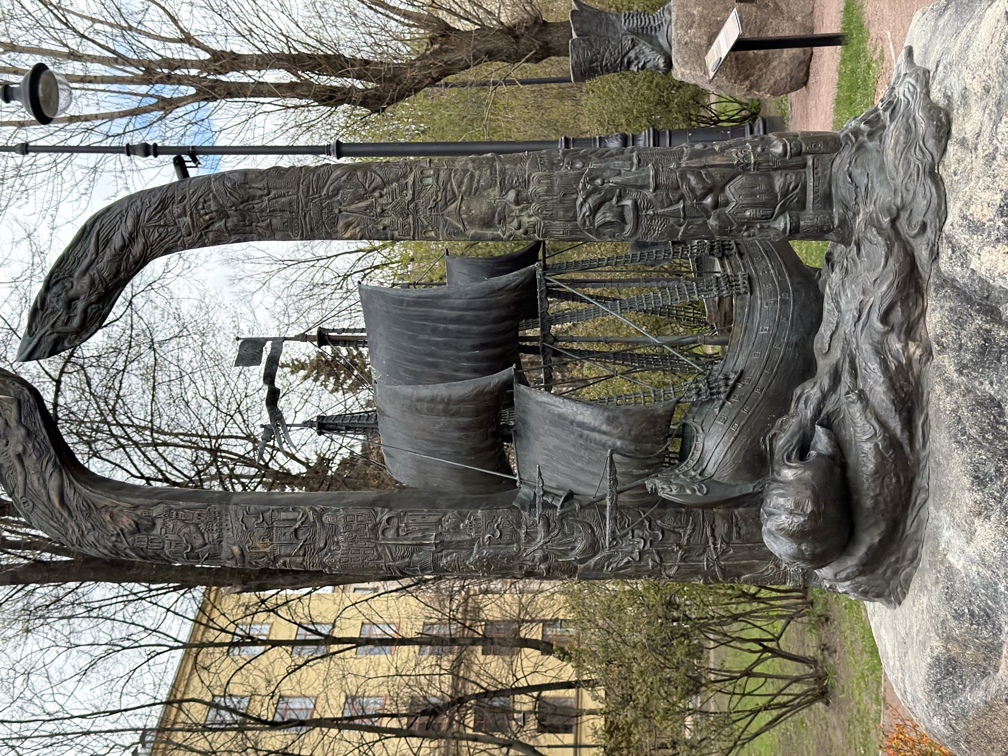 The image presents a large, dark metal sculpture, likely bronze, situated outdoors in Saint Petersburg, Russia, under an overcast daytime sky. The central feature of the artwork is a detailed historic sailing ship, possibly a galleon, complete with multiple masts and dark, flowing sails, appearing as if navigating through tumultuous waters.
The ship is integrated into a larger sculptural composition, flanked by two towering, curved structures that resemble stylized columns or massive tusks. These structures are extensively adorned with intricate relief carvings depicting a variety of scenes, including figures, buildings, and elements suggestive of historical or mythological narratives. At the base of the sculpture, near the ship's front, a prominent relief of a face, possibly human or mythical, emerges from a mass of swirling, wave-like forms, evoking a connection to the sea or a sea creature. The entire sculpture rests upon large, natural rock formations that serve as its foundation.
The surrounding environment depicts a park-like setting with numerous deciduous trees, many still with bare branches, suggesting a late winter or early spring season. Patches of green grass are visible on the ground. In the background, a light-colored building with multiple windows can be seen. A lamppost stands to the left of the sculpture. A small informational plaque on a stand is present near one of the rocks, though its text is illegible. The sculpture's detailed craftsmanship and thematic elements evoke a sense of history and maritime adventure.