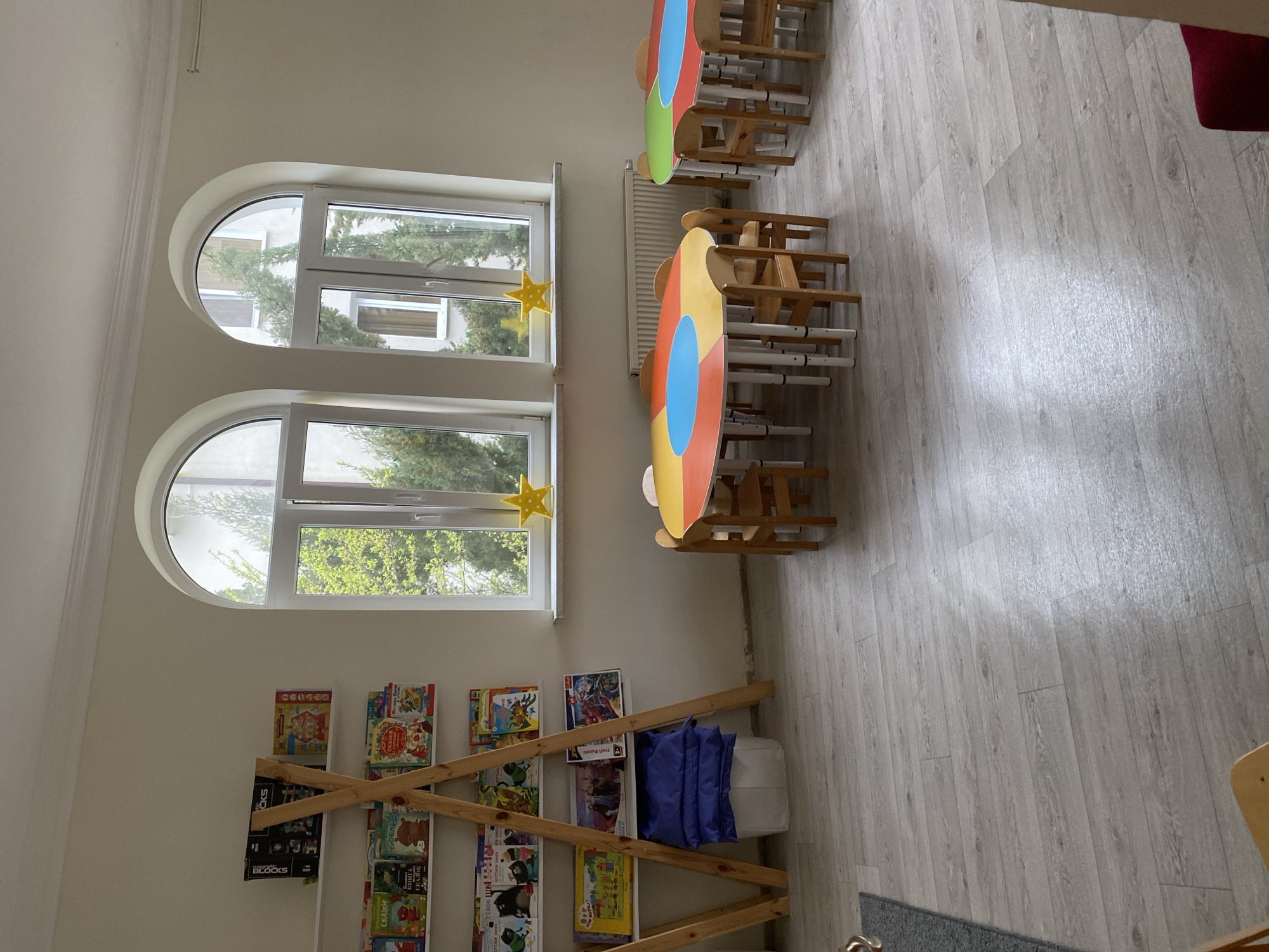 A brightly lit room with two arched windows, each adorned with a yellow star, overlooks trees outside, suggesting a natural setting. Below the windows is a bookshelf constructed from light-colored wood and filled with children’s books, blue cushions, and a white storage cube. The room's decor indicates a children's play area or classroom. Several round tables with colorful designs and wooden chairs are arranged in the room's center. The floor is light gray and appears to be made of wood or laminate. The location is specified as Sochi, Russia, although there are no distinctive landmarks or features visible in the image to confirm this claim.