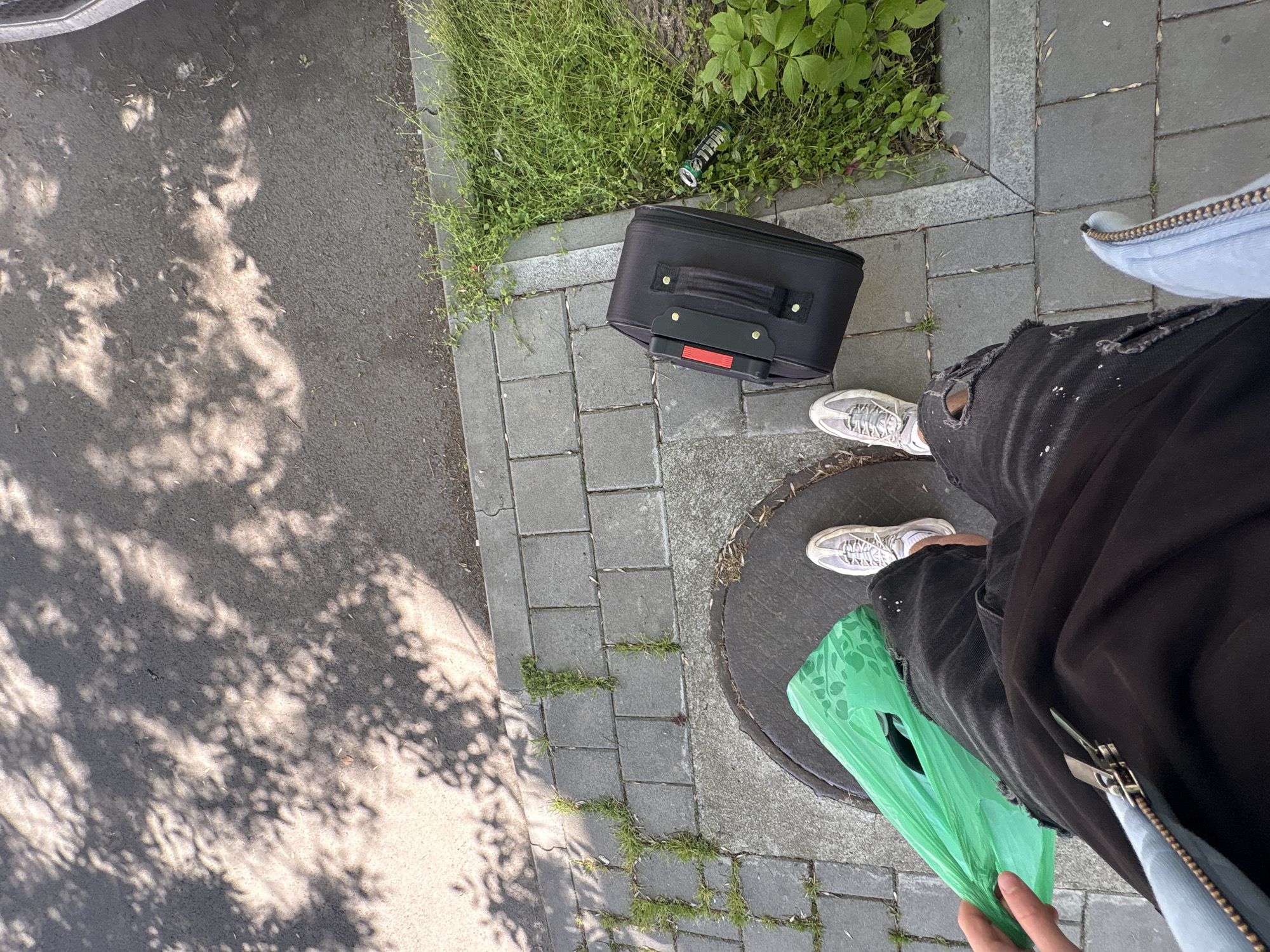 A ground-level, overhead view captures a scene on a paved sidewalk in Plovdiv, Bulgaria.
To the left, the sidewalk surface is partially obscured by tree shadows. Adjacent to the paved area, there's a patch of green grass and various plants.
A black, upright suitcase with a red handle extension lies on the pavement, near a discarded beer can nestled in the grass.
A person, seen from the torso down, stands on the sidewalk. They wear ripped black jeans and white sneakers. They are holding a green plastic bag. Part of a light blue denim jacket is visible. The person's hand is slightly visible at the bottom of the frame. The person is standing on a manhole cover on the right part of the image.