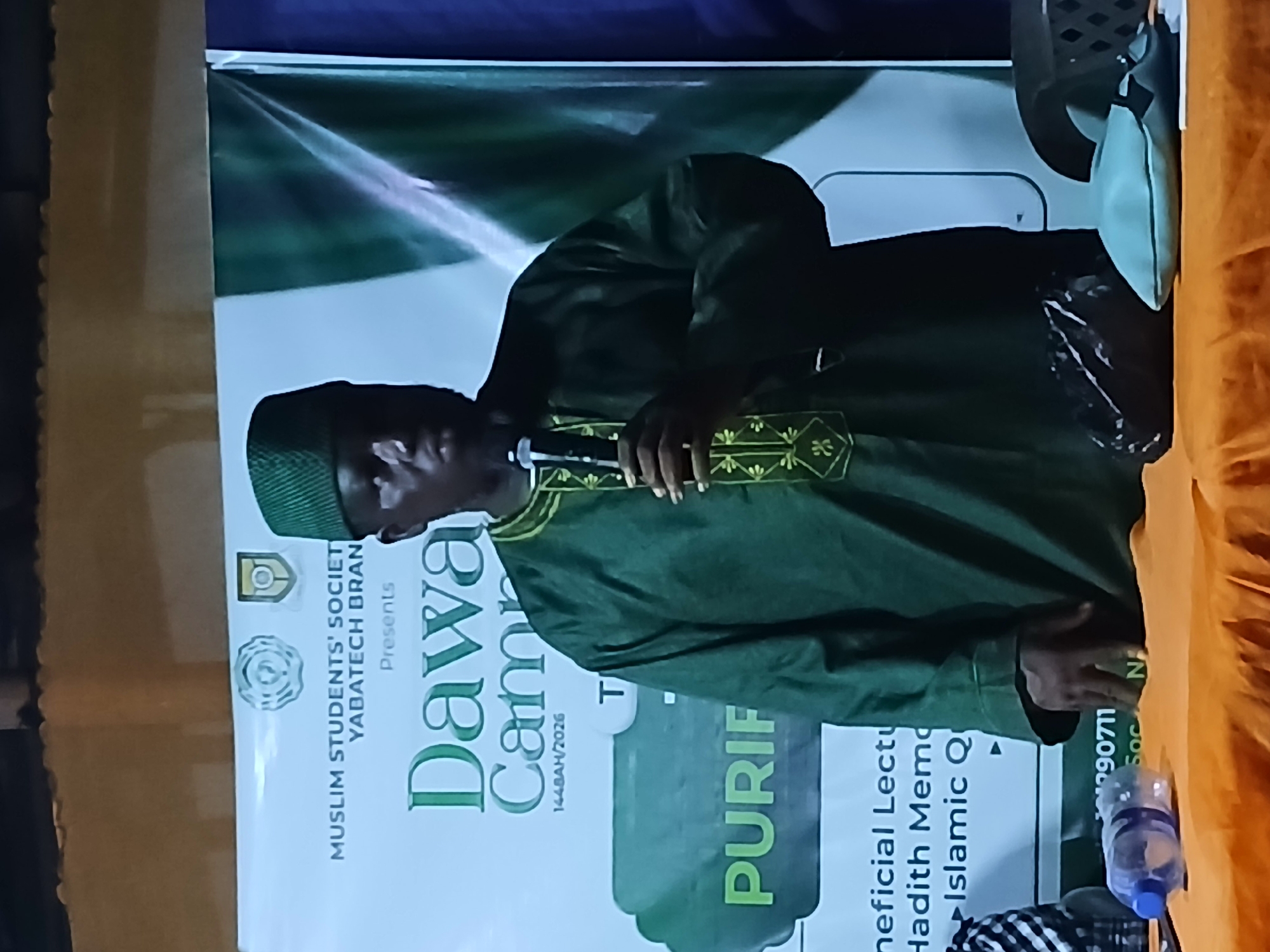 An individual wearing a dark green traditional Islamic robe and a matching cap stands behind a table, holding a microphone. The person appears to be addressing an audience, though the audience is not visible in the frame. Behind the individual is a banner with text that reads "MUSLIM STUDENTS' SOCIETY YABATECH BRANCH Presents Dawa Camp 1448AH/2026" and "PURIF". A water bottle and some papers are on the table in the foreground. The scene is lit by artificial lights, creating a stage-like atmosphere. The background suggests an indoor or semi-outdoor setting with a textured wall.