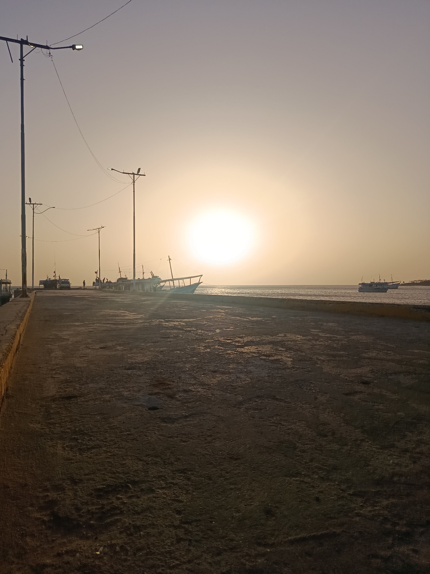 This image shows a paved pier in Juan Griego, Venezuela, at dusk or dawn. The sun is low on the horizon, creating a bright, hazy light.
Several fishing boats are docked along the pier. Some have people on or near them. Streetlights line the pier. The pier's edge has a low, yellow-tinged border. Water stretches towards the horizon with another boat visible in the distance. The pier surface is cracked and worn.