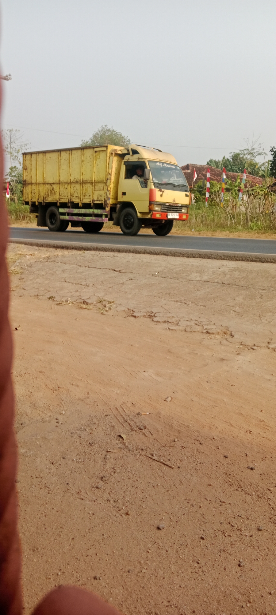 A yellow, box-style truck with a multi-colored stripe on its side is driving on an asphalt road. The truck has dual rear wheels. The driver is visible inside the cab. The truck is traveling from left to right in the image. Behind the truck, there are some trees, houses with red roofs, and flags. The foreground of the image is a dusty, dirt road with some cracks and dry vegetation. The sky is overcast. The location is Sidodadi, Indonesia.