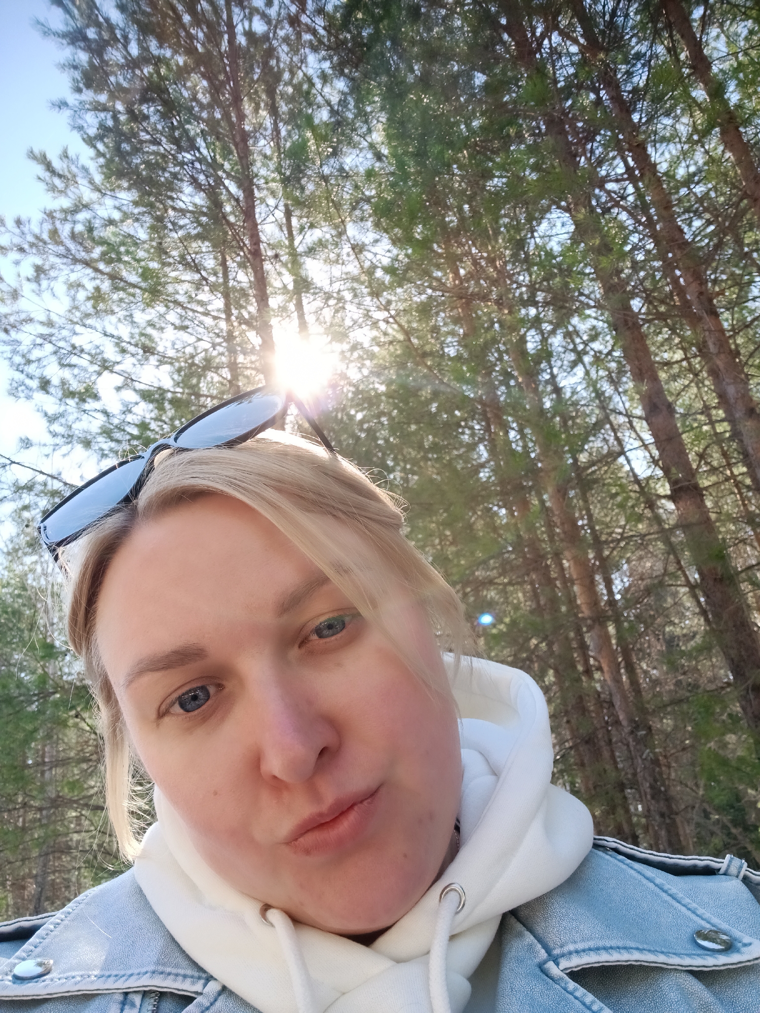 A selfie of a blonde woman with blue eyes is taken outdoors. She wears sunglasses on her head, a white hoodie, and a light blue denim jacket. The background shows a forest of tall, thin trees with sunlight filtering through the branches. This location is in Chaikovsky, Russia.