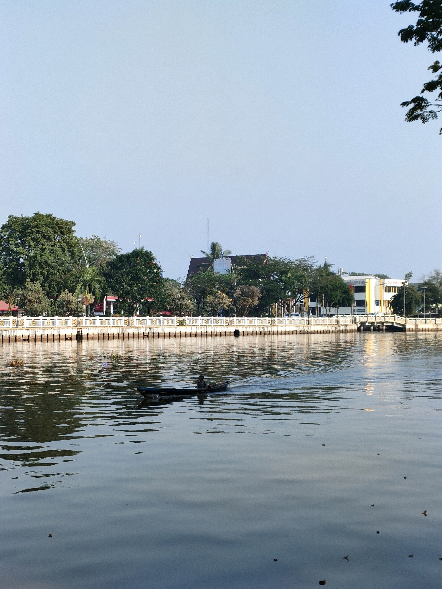 A serene, wide shot captures the Martapura River in Banjarmasin, Indonesia, bathed in the soft light of a clear, pale blue sky. The calm water reflects the environment, with gentle ripples spreading from a small, dark boat navigating across the frame from left to right. A single individual is seated in the boat, seemingly operating it, as evidenced by the wake trailing behind.
Along the riverbank, a decorative, yellow-capped white railing delineates a walkway lined with lush green trees, including a prominent palm tree. Beyond the trees, various buildings are visible, including a multi-story building with yellow accents and a darker, roofed structure. The overall impression is one of tranquility and daily life along the river.