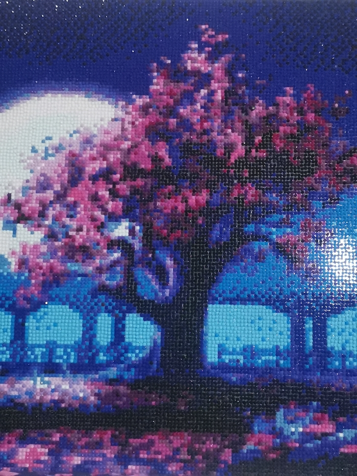 This image is a diamond painting depicting a stylized nocturnal landscape set in Старая Преображенка, Russia. A large tree with a dense canopy of vibrant pink and magenta foliage occupies the central and right portions of the composition. A bright, full moon, rendered in white and light blue, is positioned in the upper left quadrant, contrasting with a dark blue sky that exhibits subtle speckling. In the midground, below the tree, several blocky, light blue rectangular structures, interpreted as buildings, are visible. The foreground features a reflective surface, likely water, displaying fragmented reflections of the sky, tree, and structures in shades of blue, purple, and pink. No human figures, specific subjects beyond the landscape elements, actions, or interactions are present. The entire artwork presents a pixelated appearance consistent with its medium.