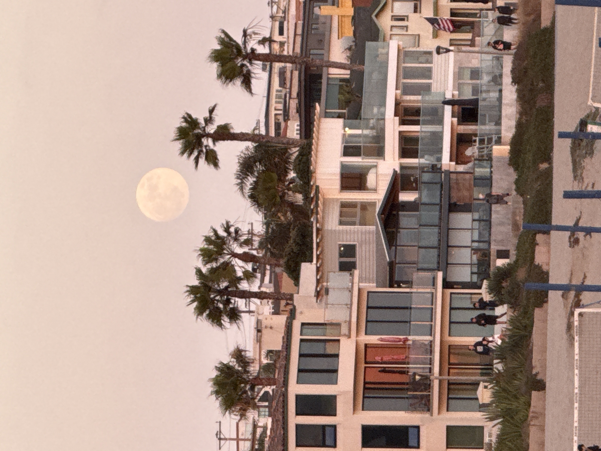 The image captures a twilight scene in Manhattan Beach, California, with a full moon prominently displayed in the pale pink sky. A cluster of multi-story beach houses, characterized by their modern architecture, glass balconies, and large windows, dominates the mid-ground. Several palm trees are scattered among the buildings, their fronds silhouetted against the sky.
In the foreground, a sandy beach area is visible, with a volleyball net and posts suggesting recreational activity. A few individuals are seen walking or standing on the elevated walkways and patios of the houses, as well as on the beach itself. One group of people is gathered on a raised area in front of one of the houses, while others are more scattered. An American flag flies from a pole on one of the buildings. The overall impression is one of a serene evening in a popular coastal community.