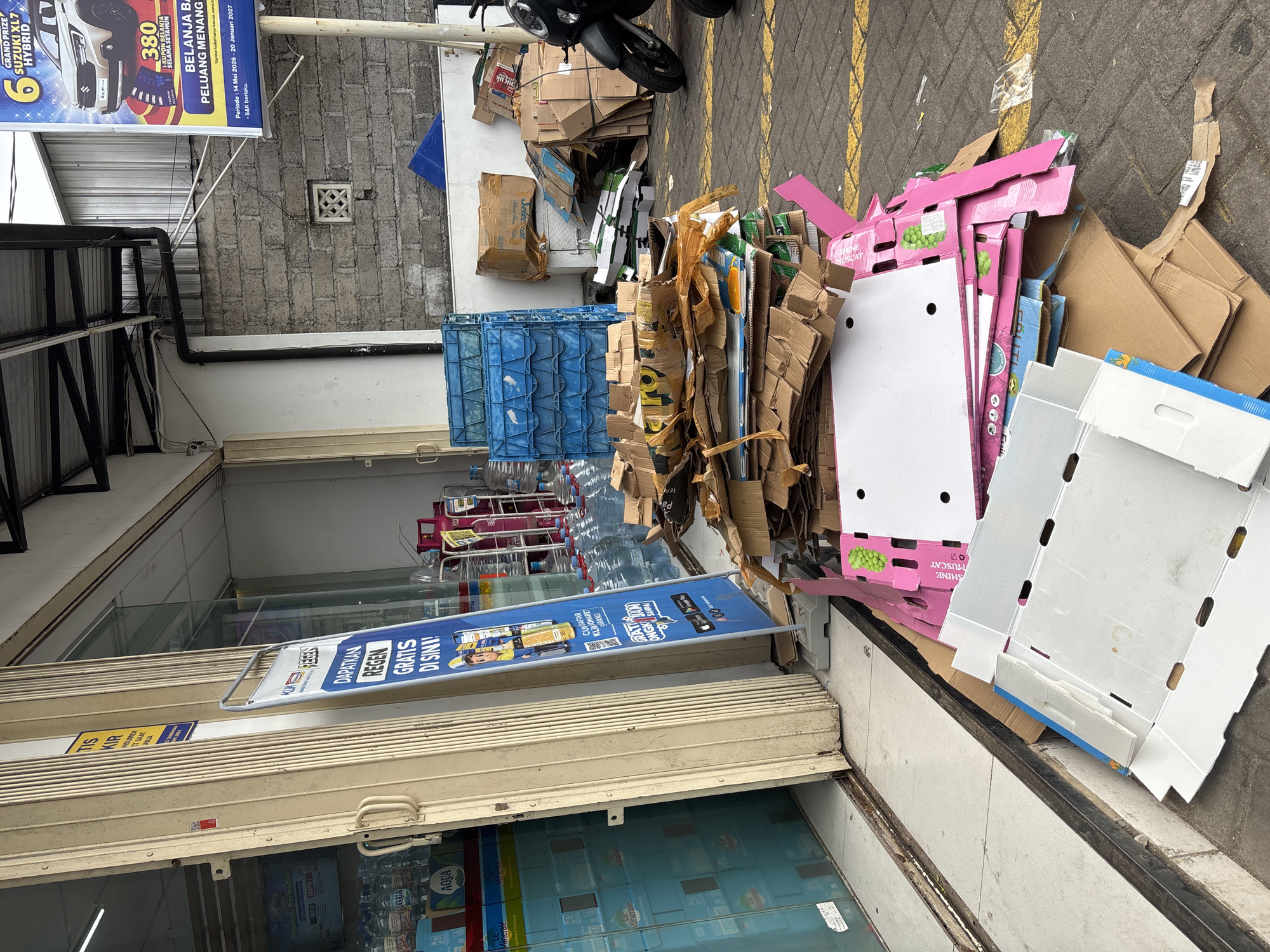 In Kuta Selatan, Indonesia, outside a store with a cream-colored metal accordion gate, a large pile of discarded cardboard boxes is visible. The boxes are various sizes and are stacked haphazardly on a paved surface marked with yellow lines. To the left of the pile, in front of the store, a blue and white promotional stand with text in Indonesian is propped up. The store's entrance appears to be a glass door. Behind the cardboard pile, several stacks of blue plastic crates are present. In the background, a section of a brick wall is visible, along with some advertising posters. A black motorcycle is parked to the right of the cardboard pile. The scene suggests a commercial area with waste materials accumulated outside a business establishment.