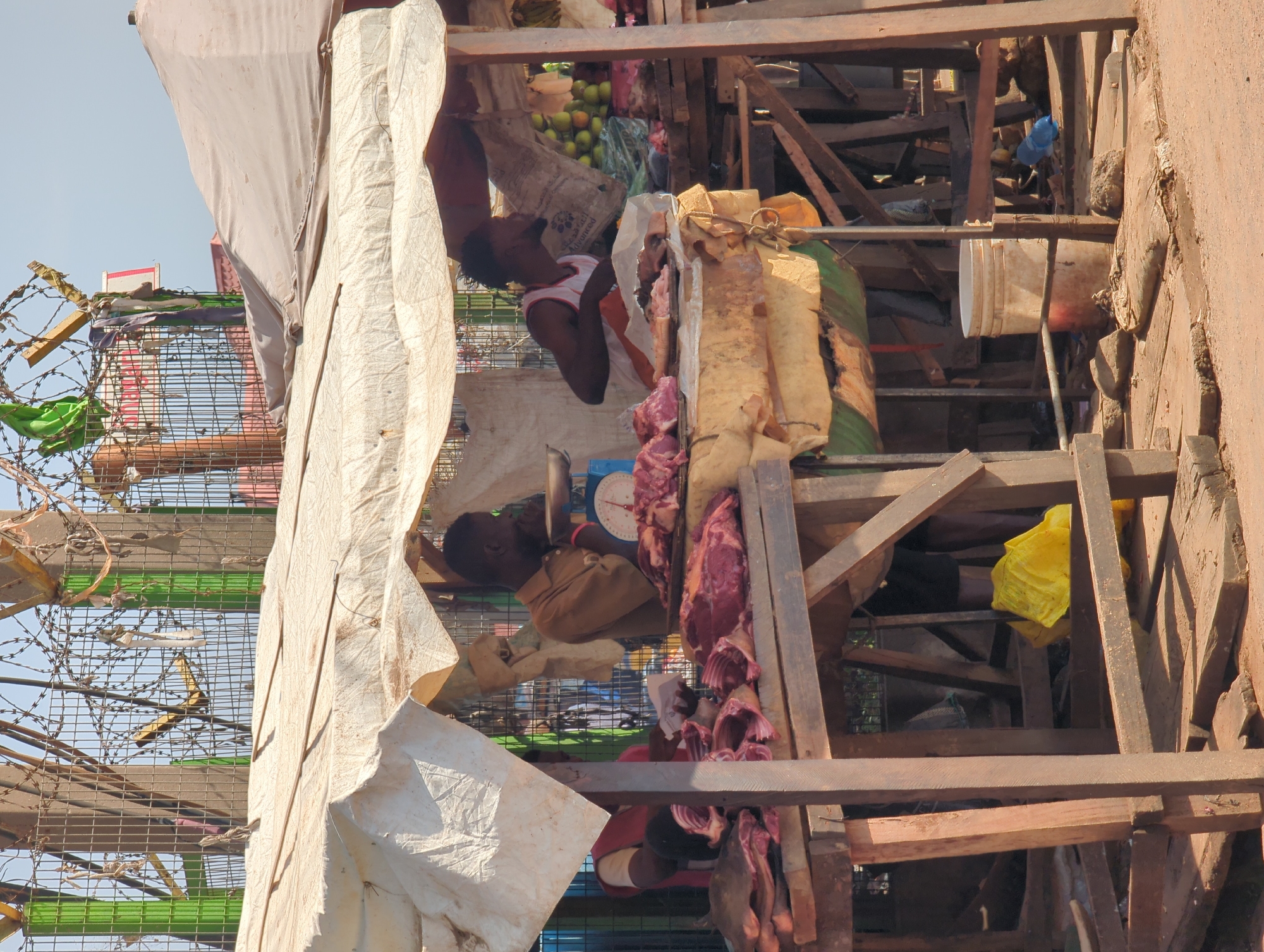 The image depicts a makeshift market stall in Narayi, Nigeria, displaying cuts of meat. The stall appears to be constructed from wooden beams and covered with white fabric. Two people are visible. One is standing inside the stall with produce, while the other is on a scale near the meat. Additional items, including bananas and a yellow plastic bag, are visible within the stall structure. Barbed wire lines the border of the stall.