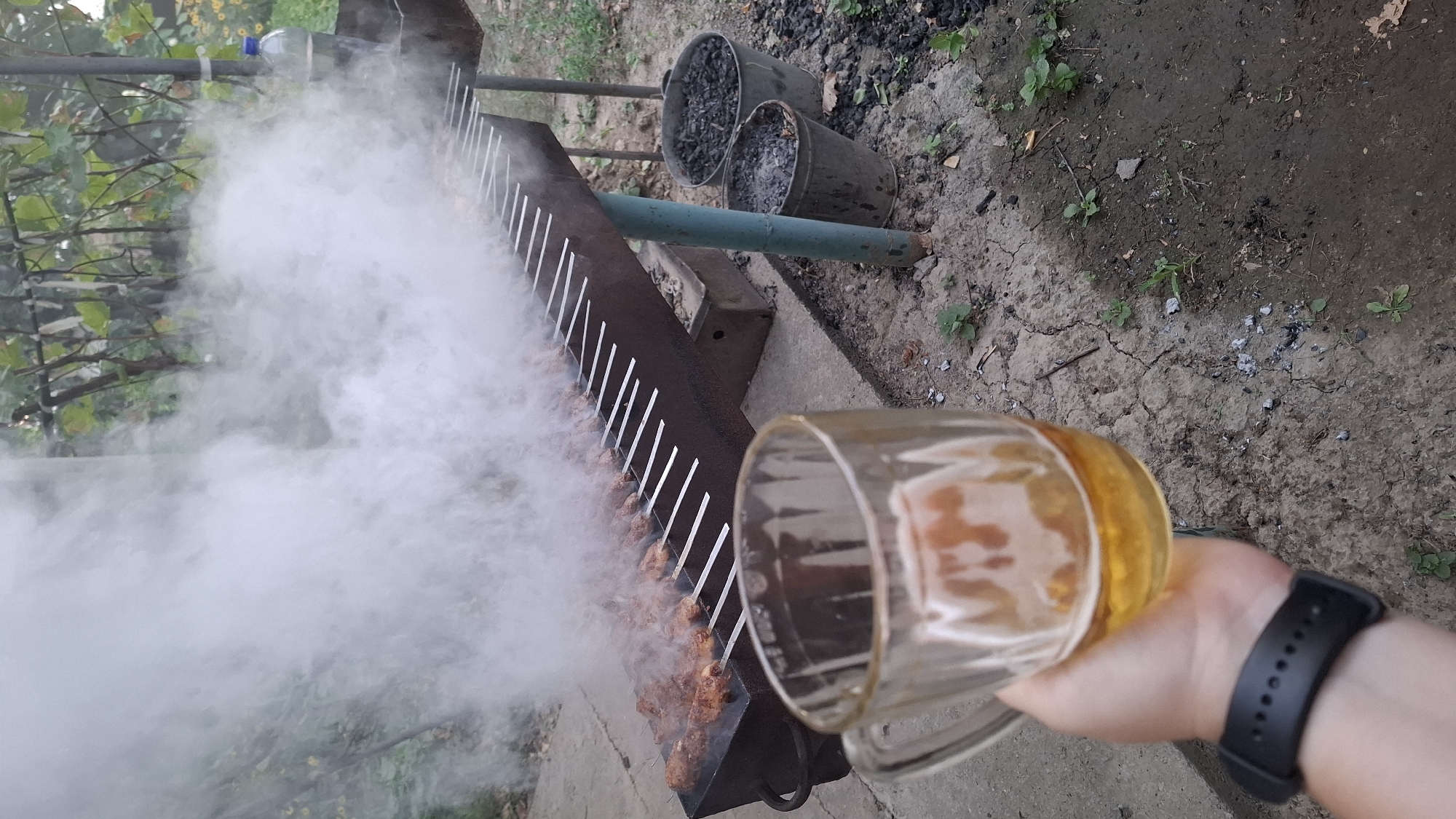 A first-person perspective shot captures a hand holding a clear glass mug filled with beer, with a black smartwatch on the wrist. In the background, a long metal grill is set up outdoors, laden with rows of meat skewers cooking over a fire that generates a significant amount of white smoke. Two metal buckets containing charcoal are visible near the grill. The scene appears to be an outdoor gathering or barbecue, likely in a rural or suburban area, given the presence of natural vegetation and rough ground. The image does not provide specific visual cues to pinpoint the location within Шұбарсу, Kazakhstan, beyond it being an outdoor setting.