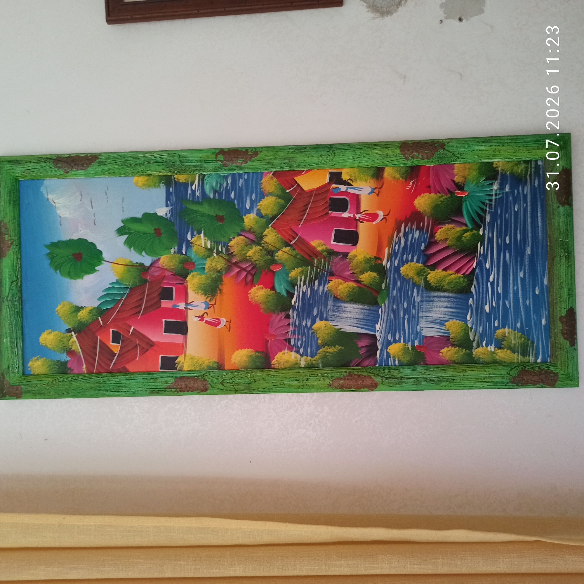 The image shows a vibrant, naive-style painting of a tropical village scene, framed in a distressed green wooden border. The painting depicts two small, brightly colored houses, one pink and one yellow, nestled amongst lush green foliage and trees. A waterfall cascades into a blue river, where two figures in traditional clothing are visible. The sky is a bright blue with some white clouds. The painting is hung on a white wall, with a yellow curtain visible on the left side of the frame. A date stamp and time "31.07.2026 11:23" are superimposed in the bottom right corner. The painting is not located in Modugno, Italy, but is a standalone artwork.