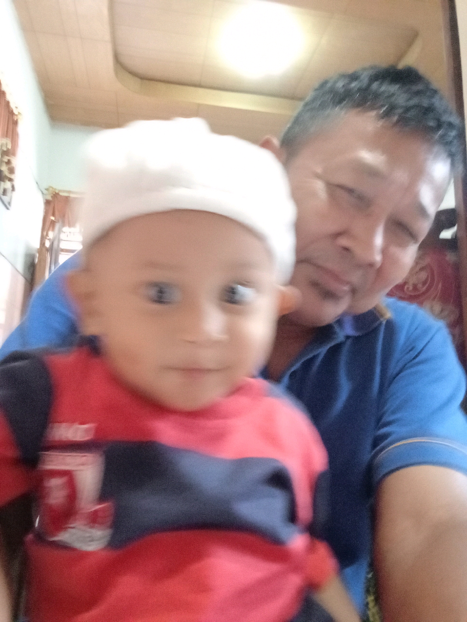 An adult male and a young child are pictured in a selfie taken indoors. The adult male, on the right, has short, dark hair and is wearing a blue collared shirt. The child, on the left, is wearing a white hat and a red shirt with blue accents. The background appears to be a room with a ceiling fan and wooden paneling. Based on context, the location is identified as Kebonagung, Indonesia.