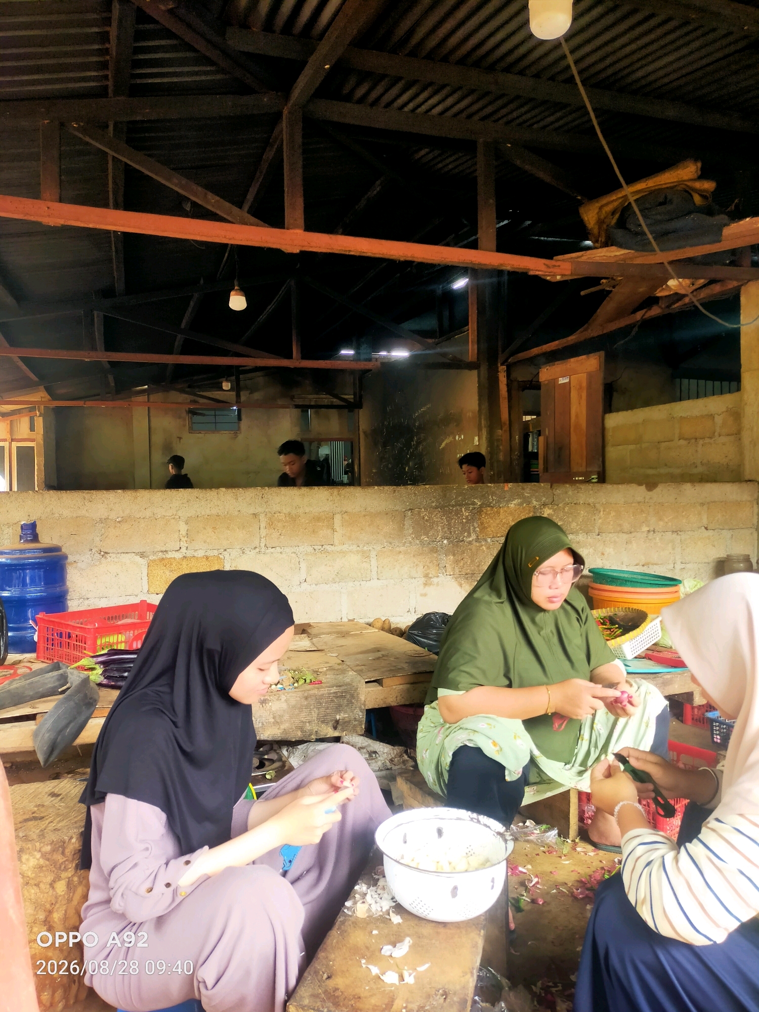 In a dimly lit, open-air structure with a corrugated metal roof supported by wooden beams, three individuals are engaged in food preparation. The setting appears to be a market or a communal kitchen, with rough concrete block walls forming partitions. In the foreground, two women are seated on the floor, diligently peeling and chopping onions. One woman, wearing a green hijab and a matching outfit, wears glasses and has her hands occupied with an onion. The other woman, in a black hijab and a light purple outfit, is also focused on her task. To the right, a third person, partially visible and wearing a light-colored hijab and a striped shirt, is also involved in the food preparation. In the background, through a gap in the wall, two young boys can be seen. The floor is littered with onion skins and other debris. A large blue water jug, red plastic crates, and various baskets containing vegetables are scattered around the area, suggesting ongoing activity. The image is timestamped "2026/08/28 09:40" and displays "OPPO A92" at the bottom left, indicating it was taken with an Oppo A92 phone. The environment is modest and functional, indicative of a working space in Suka Maju, Indonesia.