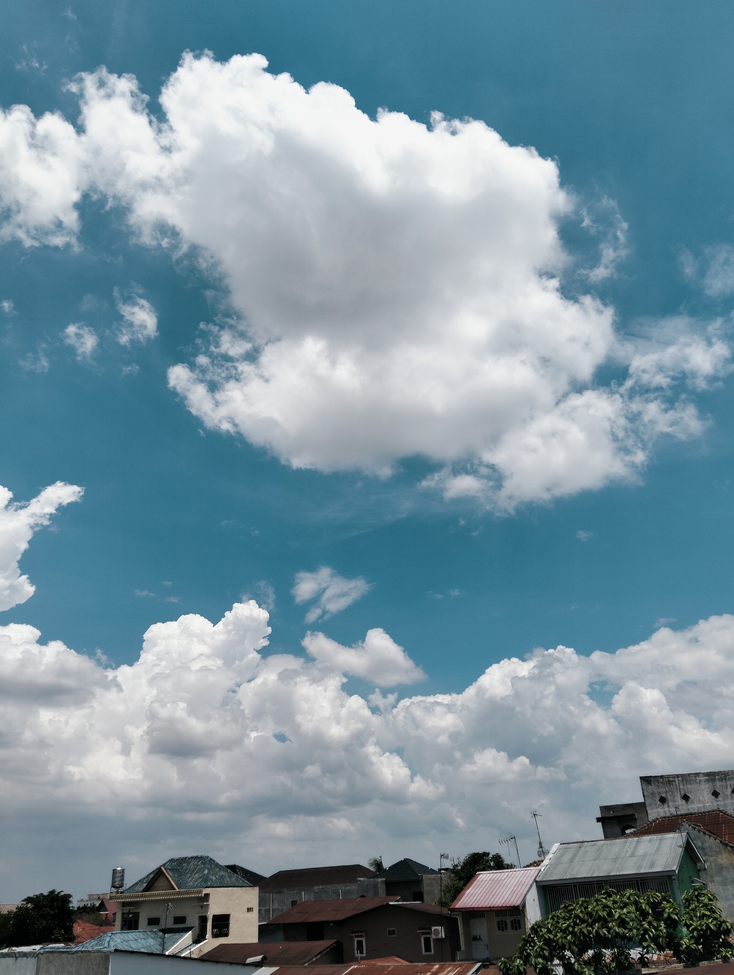A photograph taken in the city of Medan, Indonesia, shows a bright blue sky filled with voluminous white clouds. In the foreground, rooftops of various buildings are visible, suggesting a residential or urban area. The architecture appears to be a mix of styles, with some buildings featuring tiled roofs and others having corrugated metal roofing. Trees are interspersed among the structures. The lighting suggests it is daytime, likely mid-morning or afternoon, given the clarity of the sky and the shadows cast. There are no people or animals clearly visible in the image. The overall scene captures a typical sky view over a city in a tropical climate.