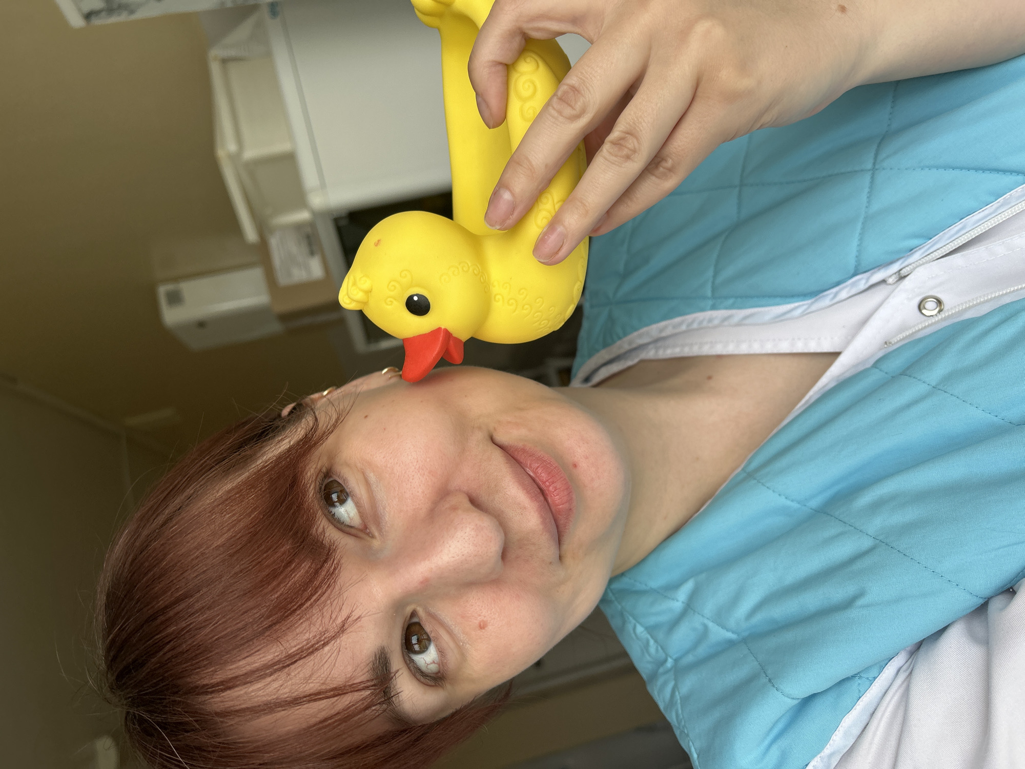 An indoor portrait shows a woman, depicted from the chest up, with reddish-brown hair and brown eyes, looking directly at the viewer with a gentle, slightly smiling expression. She is wearing a light blue quilted jacket, which has a white zippered front and white trim, over a white garment. Her right hand is holding a bright yellow rubber duck toy, which has black eyes and a reddish-orange beak, pressed against her right cheek. The duck features subtle embossed swirl patterns on its body. The background is an indoor setting with light-colored walls and indistinct white objects, possibly furniture or architectural elements, suggesting a room or office environment. The overall mood conveyed is pleasant and lighthearted. No visible text is present in the image. The photo was taken in Фоки, Russia.