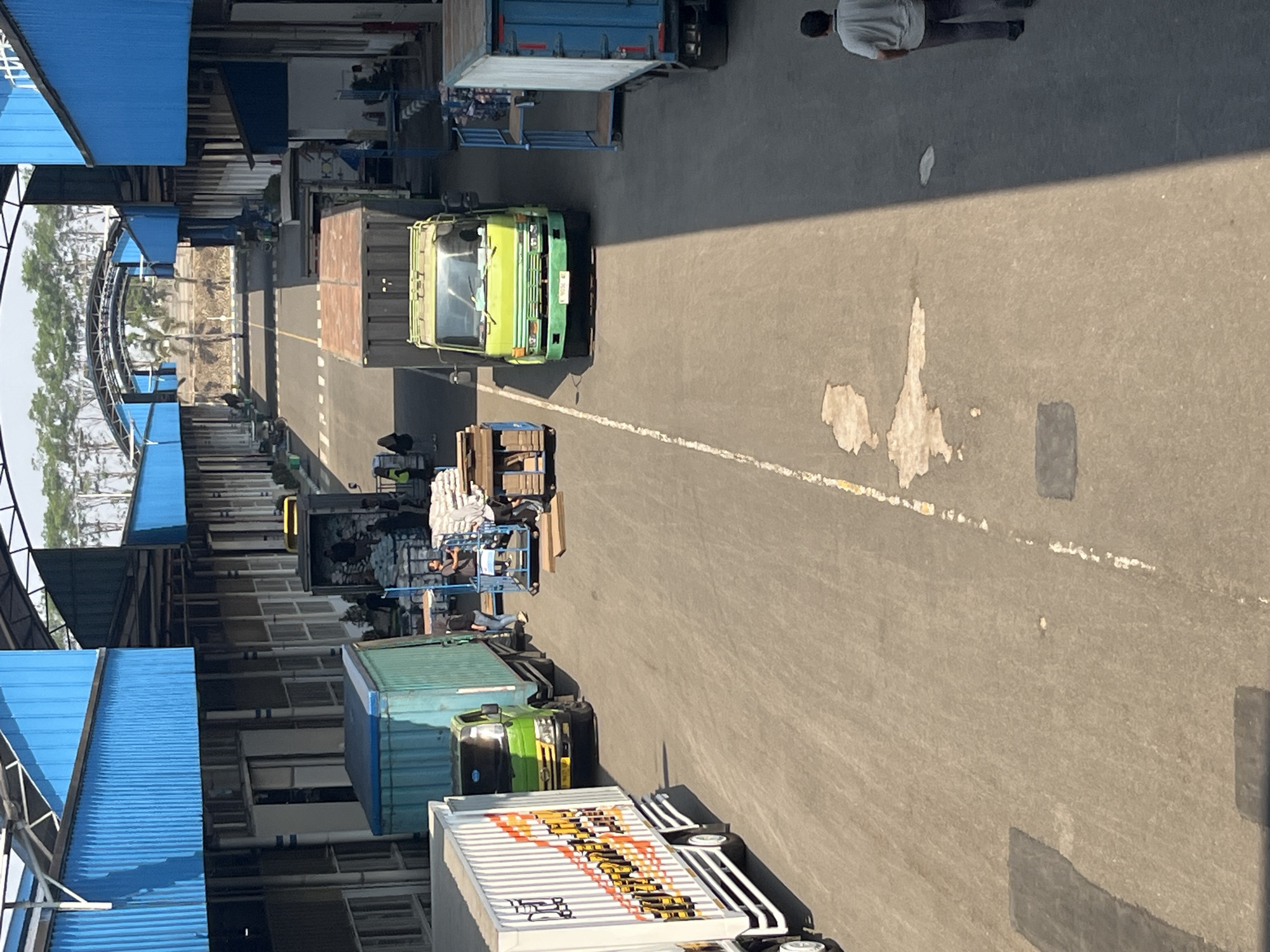 The image depicts an outdoor loading bay area at what appears to be a logistics or distribution center in Pelang, Indonesia. The scene is characterized by a long, open-air structure with bright blue corrugated metal roofing supported by metal beams. Several trucks are parked or in motion within the loading bay.
On the left side of the frame, a white truck with a red and yellow graphic design is partially visible. Next to it is a green truck with a turquoise cargo container. Further down the lane, a bright green truck is positioned, facing away from the camera. To its right, a blue cargo container truck is parked.
Several individuals are present in the scene, engaged in activities related to cargo handling. Near the green truck with the turquoise container, a few people are gathered around a blue trolley loaded with sacks, seemingly in the process of moving or inspecting goods. Another individual, wearing a grey t-shirt and dark trousers, walks away from the camera in the lower right portion of the image. In the distance, at the end of the loading bay, an arched structure is visible, possibly an entrance or exit to the facility, with trees and a dry, dusty landscape beyond. The ground is paved asphalt with white and yellow painted lane markers and some irregular lighter-colored patches, possibly oil stains or dried residue. The sunlight casts distinct shadows, indicating it is likely daytime.