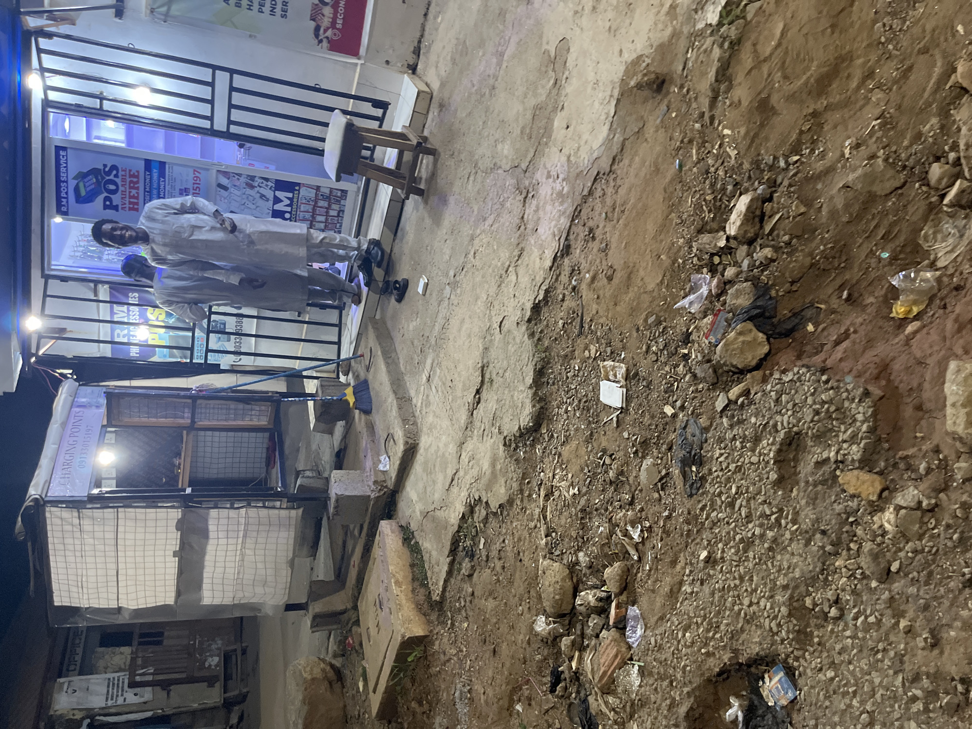 The image depicts a street scene in Abuja, Nigeria, at night. The foreground is dominated by rough, uneven terrain with loose rocks, dirt, and scattered debris, including plastic bags and broken bricks, suggesting an unpaved or damaged area. In the mid-ground, a paved walkway or street is visible, also showing signs of wear and tear, with a concrete slab on the left and a broken section of pavement.
On the right side of the mid-ground, a small, open-fronted shop or service stall is present. It has a metal frame and is covered with a white checkered fabric. Inside, a light illuminates the space, and shelves are visible, holding various items. A sign above the stall reads "CHARGING POINTS" with a phone number below it. Adjacent to this stall, another business is visible, with a more structured storefront featuring glass doors and interior lighting. A sign inside this business reads "R.M POS SERVICE" and prominently displays "POS AVAILABLE HERE" along with information about money services. Two men are standing in front of this POS service, one facing the camera and smiling, dressed in traditional Nigerian attire. The other man is partially visible, facing away from the camera. A simple wooden stool with a padded seat is placed in front of the POS service. A broom with a yellow handle and blue bristles stands near the "CHARGING POINTS" stall. The overall environment appears to be a bustling but somewhat unkempt commercial area within the city.