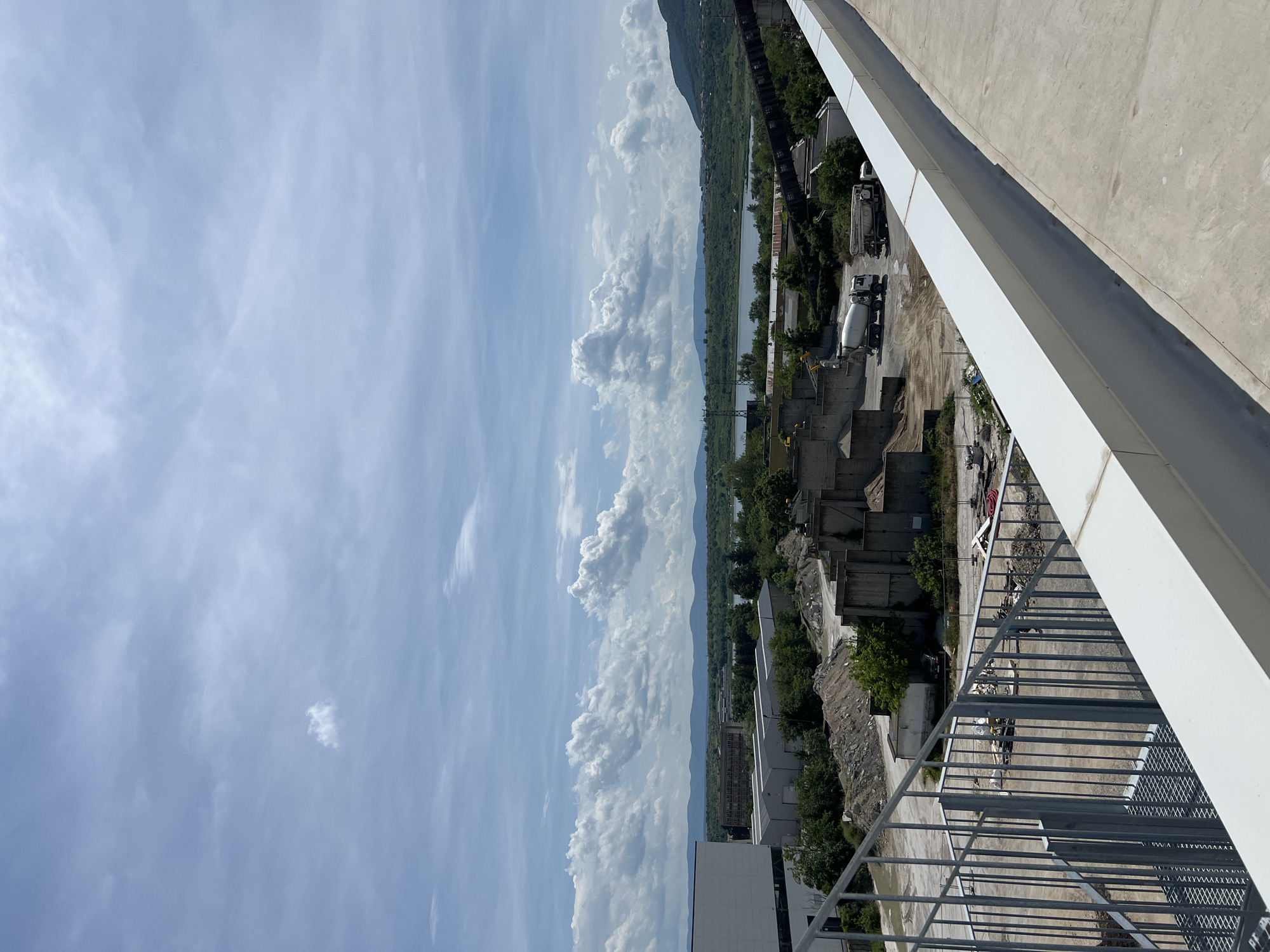 An outdoor, high-angle view captures a landscape dominated by a bright blue sky with scattered white cumulus clouds. The image is oriented vertically, with the sky filling the left half. Towards the middle of the frame, a distant body of water, likely a river or lake, is visible, bordered by a long stretch of green land and distant mountains or hills.
Below this natural horizon lies an industrial or construction area. This section features several concrete structures, piles of aggregates or debris, and patches of green trees. Multiple large white concrete mixer trucks are parked within this zone, suggesting ongoing or recent construction work.
The right side of the image is dominated by architectural elements of the vantage point. A clean, modern white structure, possibly a building facade or a bridge beam, cuts diagonally across the upper right corner. Below this, a grey metal safety railing, composed of horizontal bars and mesh panels, extends along the lower right, indicating a balcony or walkway from which the photograph was taken.
The scene appears to be well-lit, indicating a clear, sunny day. No human figures or discernible text are visible.