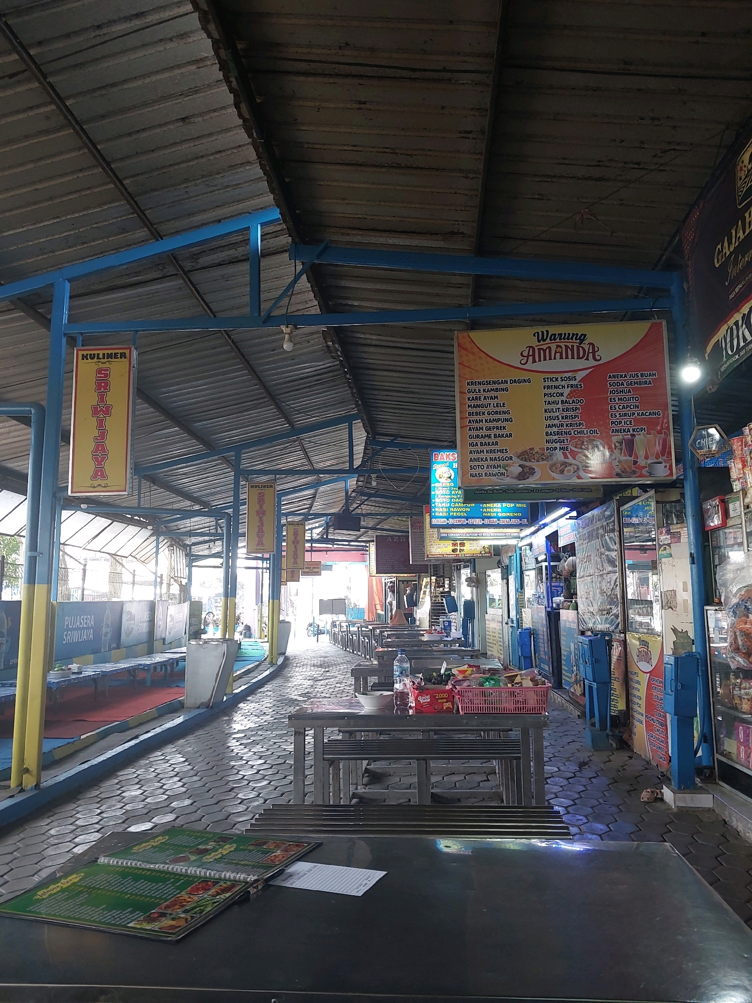 The image depicts an outdoor food court or street food alley, likely in Malang, Indonesia. The scene is characterized by a covered walkway with a corrugated metal roof supported by blue metal beams and poles. The ground is paved with hexagonal gray tiles. On the left, there are long, blue-topped tables and benches, some of which are set up for dining. Yellow poles with "KULINER SRIWIJAYA" signs are visible. Further down the alley, numerous metal tables with chairs are arranged in rows, indicating a seating area for customers.
A prominent sign for "Warung Amanda" is displayed, listing various food and drink items such as "Kremsengan Daging," "Gule Kambing," "Stick Sosis," "French Fries," "Es Jus Buah," and "Es Mojito." Adjacent to this is a stand or small shop stocked with various goods, including bottled drinks and snacks. Another sign, partially visible on the right, reads "GAJAH."
On the tables in the foreground, there is a menu, open and displaying colorful food images, as well as a white piece of paper, possibly an order slip. Several plastic baskets filled with food items and drinks are placed on some of the tables. The overall atmosphere suggests a busy but somewhat weathered commercial area. The lighting indicates it is daytime, with sunlight filtering in from the open sides of the covered area.