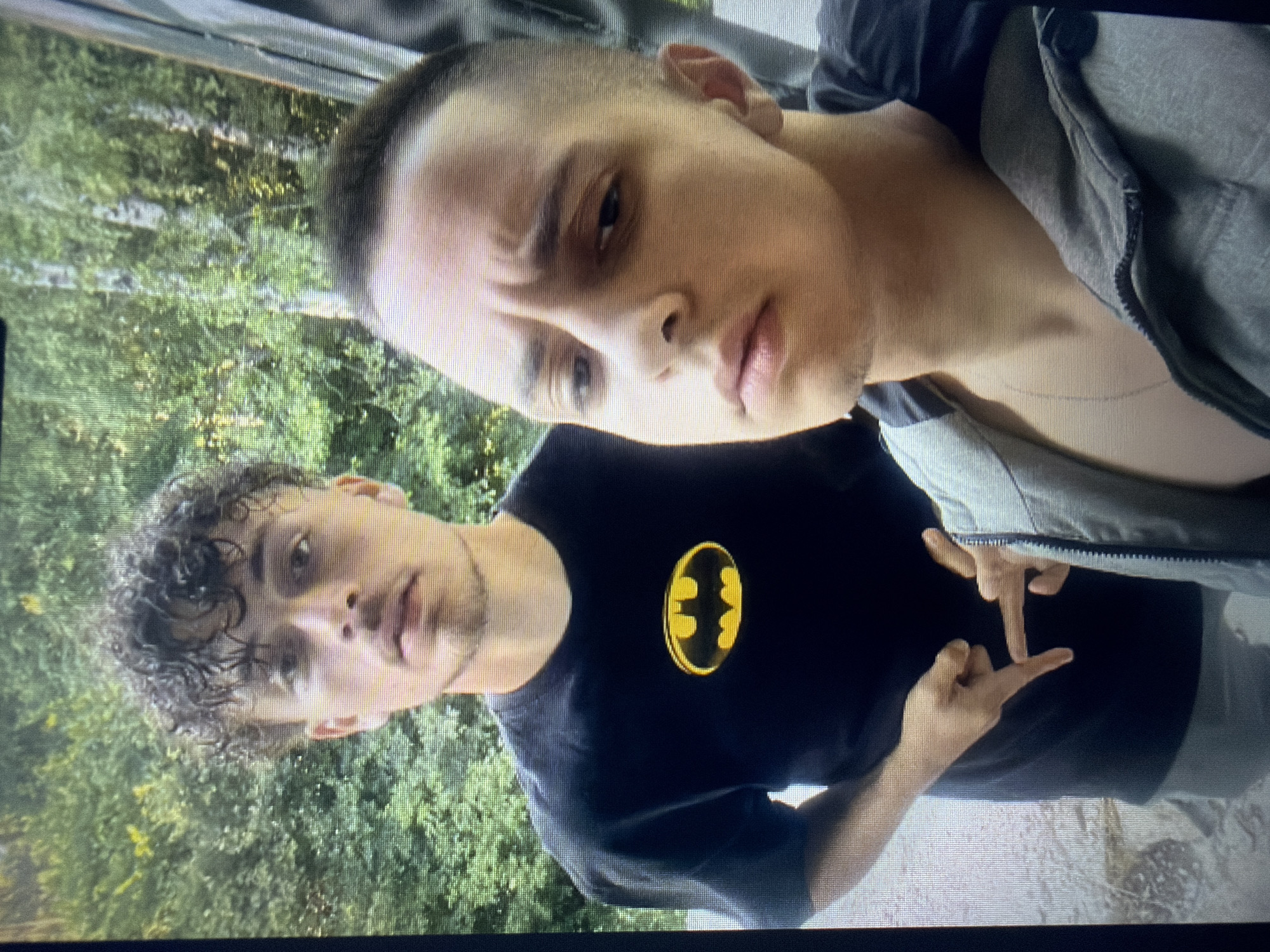 The image shows two young men in an outdoor setting. The first man, positioned in the background, has dark, curly hair. He is wearing a black T-shirt featuring the yellow Batman logo. He is making a hand gesture with both hands. The second man, in the foreground, has short, dark hair and is wearing a gray zip-up hoodie over a dark shirt. The background appears to be a wooded area, possibly a park, with green foliage visible. The location is in the city of Unknown, Unknown.
