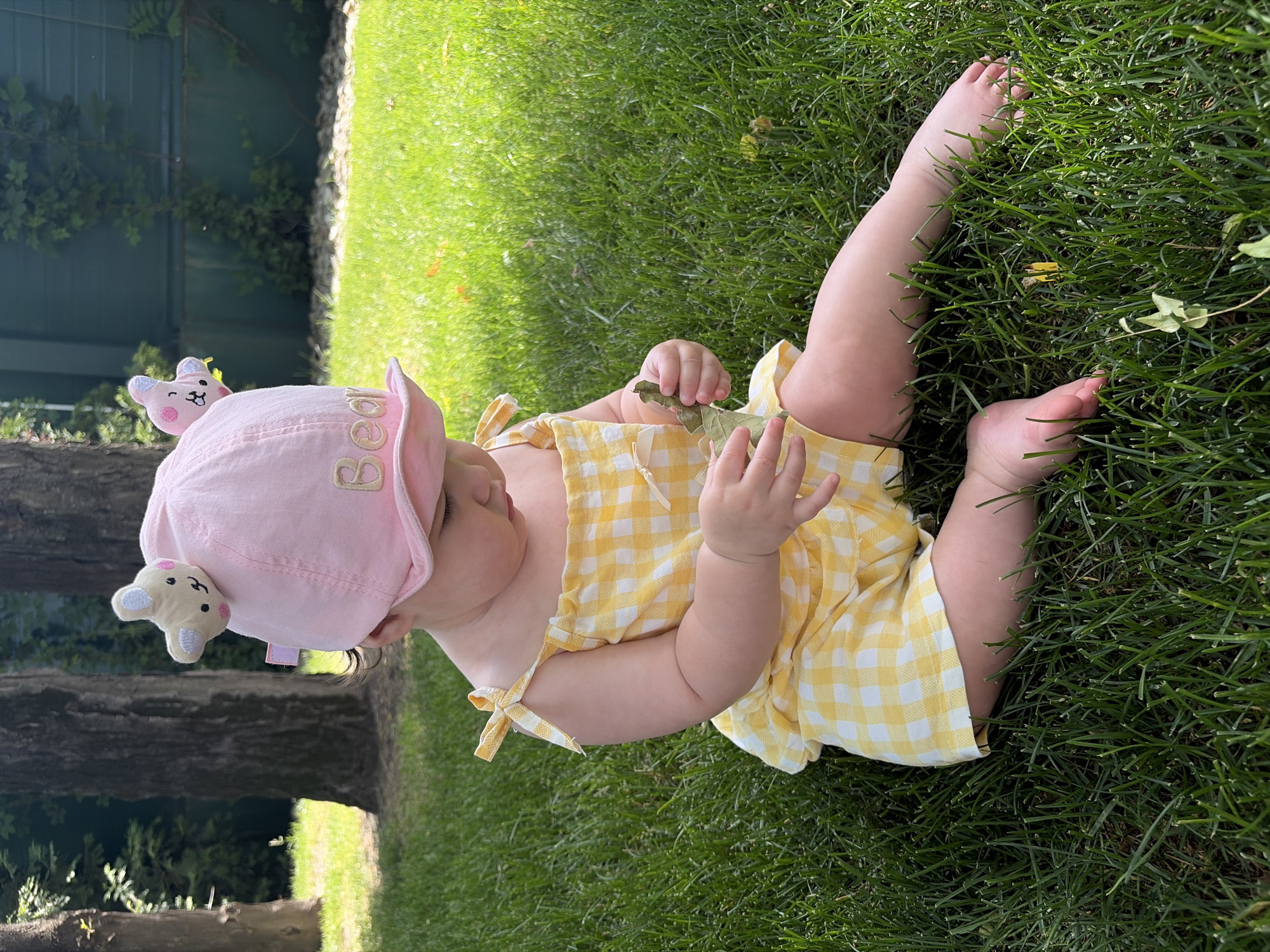 An infant is seated on a vibrant green lawn in Krasnodar, Russia, under bright daylight. The infant wears a light pink bucket hat featuring two plush, cartoonish animal ear appliques (one resembling a bear, the other a rabbit) on the crown, and the word "Bear" embroidered in gold on the front brim. A yellow and white gingham romper with tied shoulder straps covers the torso and legs. The infant's head is tilted downwards, focusing attention on a small, dried leaf or twig held in both hands. In the background to the left, multiple large trees with dark trunks and green foliage are visible. To the right, a dark green fence or wall is partially obscured by climbing green vegetation.