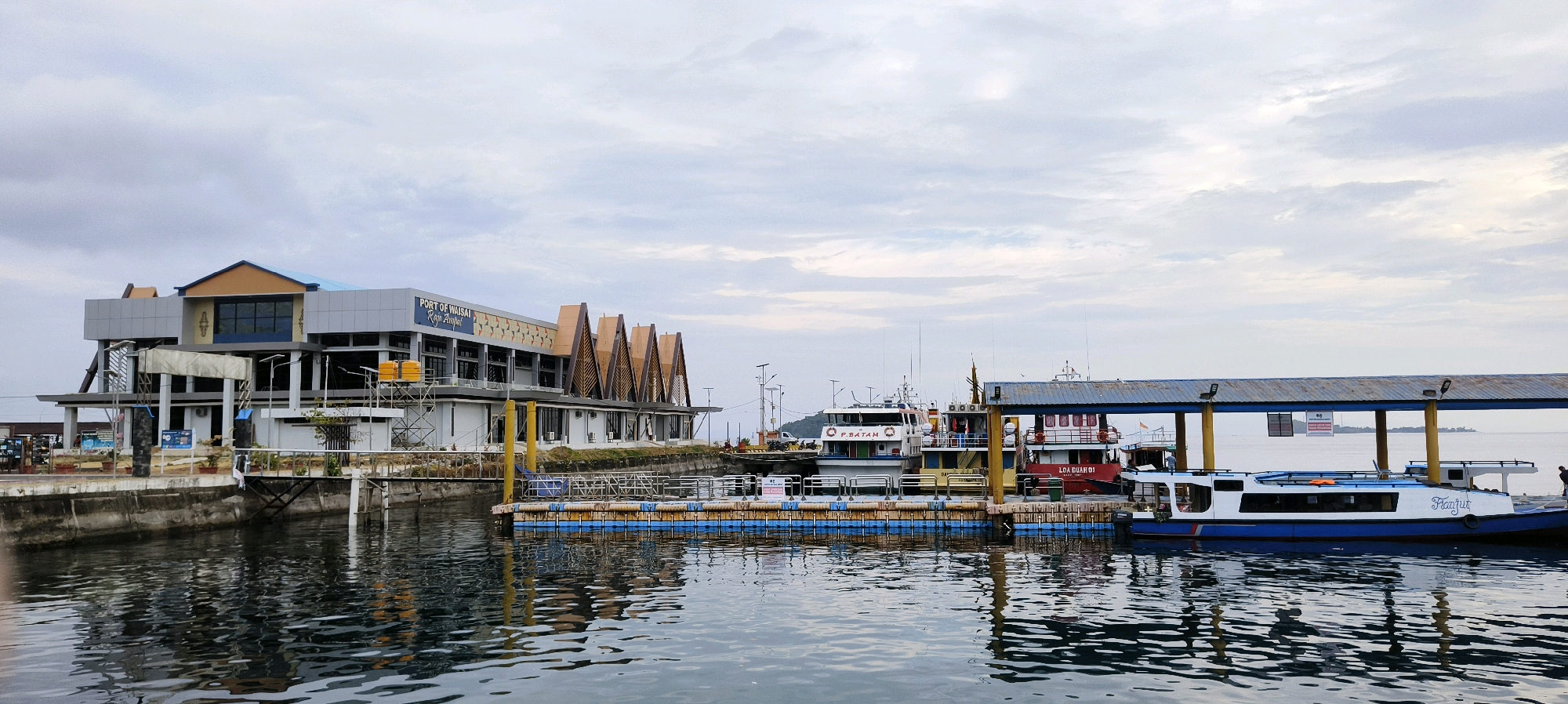 The image depicts the Port of Waisai, Raja Ampat, Indonesia, under an overcast sky. The port features a modern, multi-story building with a blue roof and gray walls, adorned with distinctive triangular architectural elements on its facade. Adjacent to the building is a floating dock with yellow and blue pontoons, where several vessels are moored. These include a white and blue passenger ferry named "Fianfur" and other boats, one with "P. BATAM" visible on its side and another labeled "LOA BUAH 01". A covered walkway extends over part of the dock, supported by yellow pillars. The water is calm, reflecting the structures and the cloudy sky. A few individuals are visible near the boats and the dock. The overall scene is that of a functional port at what appears to be an early morning or late afternoon, given the soft lighting.