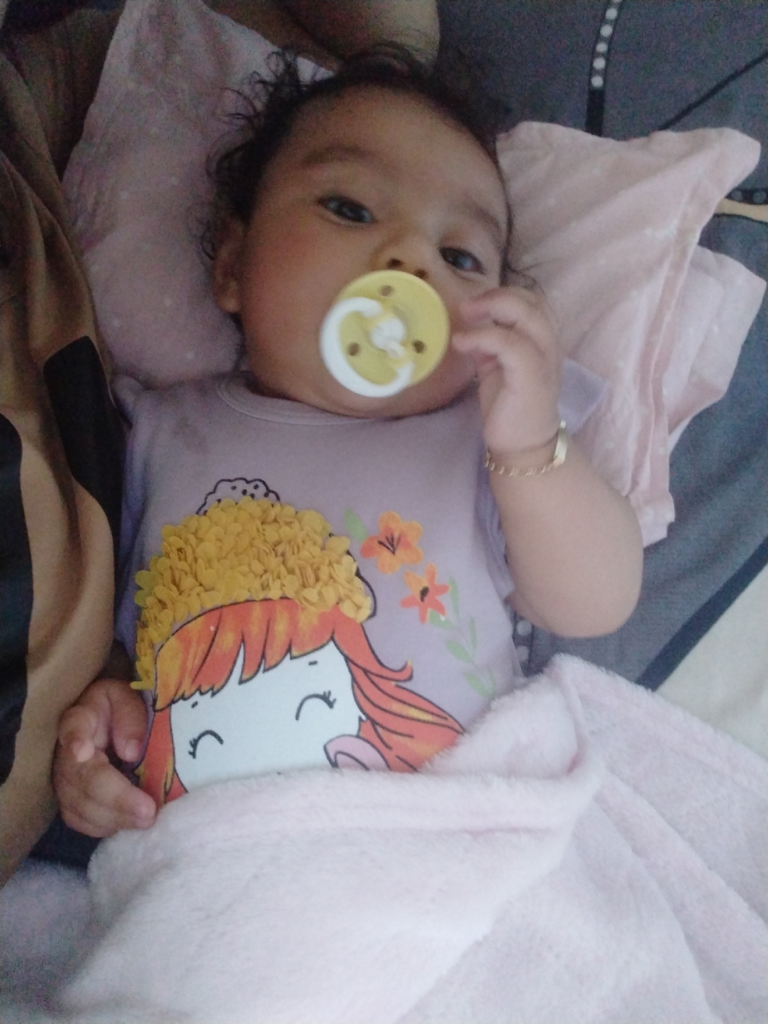 A baby is lying on a bed with a pink blanket partially covering their body. The baby has a pacifier in their mouth and is wearing a lavender shirt with a cartoon image of a girl with yellow flowers for hair. The baby is wearing a gold bracelet on their right wrist. The bedding includes pink and grey patterned fabrics. The location is Unknown, Unknown.