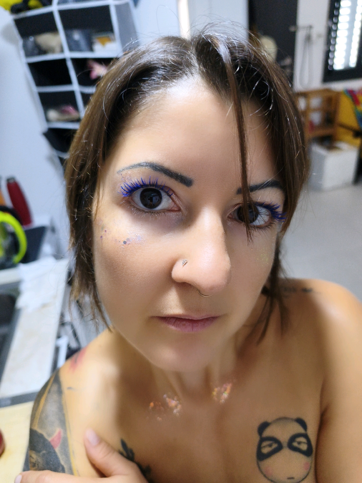 The image is a close-up, waist-up shot of a young woman with dark brown hair, dark eyes, and light skin, taken indoors. She is looking directly at the camera. The woman is wearing blue mascara and has glitter applied to her cheekbones and collarbone. She has a nose ring and a diamond stud in her right nostril. On her left shoulder and chest, there is a tattoo of a panda. She has another tattoo on her left arm and shoulder with what appear to be demonic figures. The background is blurry and includes shelves filled with various items, a window with blinds, and other household objects. The lighting suggests an indoor setting, possibly a home. The woman appears to be nude from the waist up. The image does not provide any specific information about the city of Lachish Regional Council, Israel, or any context that would place the subject there.