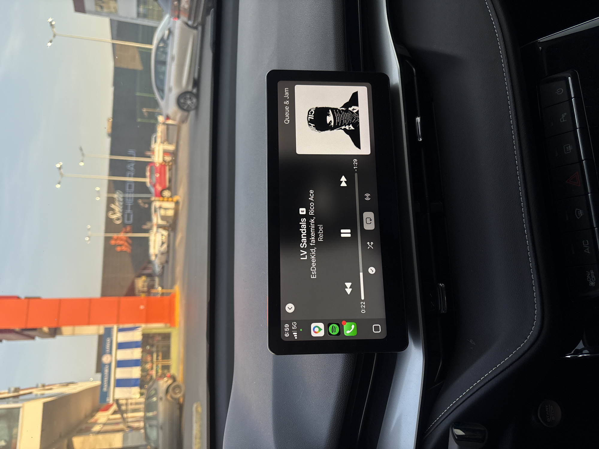 The image captures the interior of a car, focusing on a vertically oriented digital infotainment screen displaying a music player, with a view of the outside world through the windshield. The screen shows the song "LV Sandals" by "EsDeeKid, fakemink, Rico Ace" from the album "Rebel," featuring a stylized black and white portrait as album art. The track is 22 seconds in, with 1 minute and 29 seconds remaining. The display also indicates the time as 6:59, a 5G connection, and shows icons for Spotify and Google Maps.
The car's dashboard, visible around the screen, is dark with white decorative stitching. Several control buttons are present, including a "START STOP" button, an "A/C" button, and controls for parking, fan, hazard lights, and rear defogger.
Outside the vehicle, a street scene unfolds under what appears to be late afternoon or early evening lighting. Buildings, streetlights, and other vehicles, including a white car and a red car, are visible. A large sign for "CHEDRAUI," a supermarket chain, is clearly seen in the background, along with a partially visible blue and white striped sign that might indicate a "PANADERIA" (bakery). The scene is set in San Bernardino Tlaxcalancingo, Mexico, indicated by the Chedraui store.